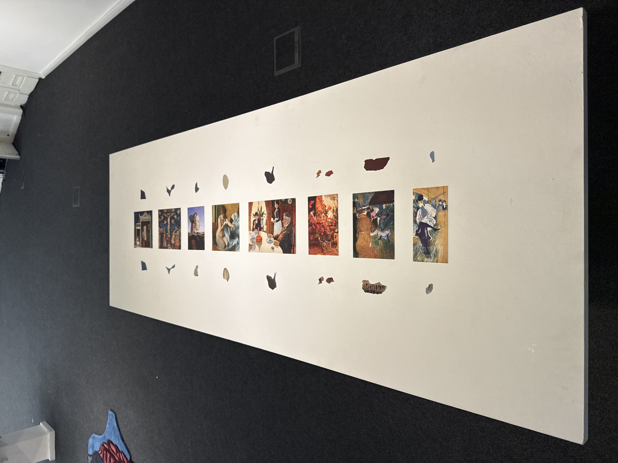 The image shows a white rectangular board affixed to a dark gray wall. The board contains a series of square images aligned horizontally, spaced unevenly. The images appear to be reproductions of paintings, depicting various scenes with people in indoor and outdoor settings. There are several irregularly shaped voids cut out of the board, positioned below the images. A dark square shape is visible in the wall above the board, possibly a ventilation opening or utility access point. To the left of the board, near the top, there's a partial view of a white ceiling with decorative molding. On the lower left, there is a multicolored rug with shapes that might resemble mountains, placed on the floor against the wall.
Without additional information, it is impossible to determine the location in Buenos Aires.