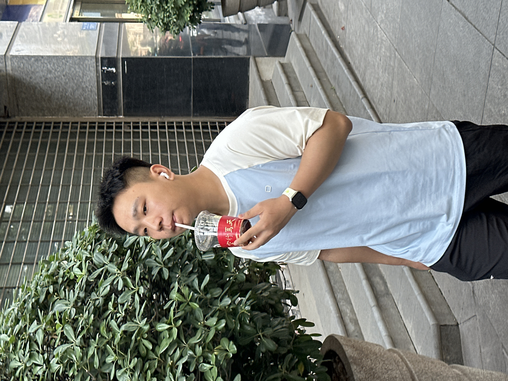 A person wearing a white and light blue t-shirt and black shorts is drinking from a clear plastic cup with a red label and a straw. They are standing outside, near a lush green bush in a planter. The background shows a building with a gray and black facade and metal grilles. Concrete stairs lead up to the building. The location is Yuzhong District, China.