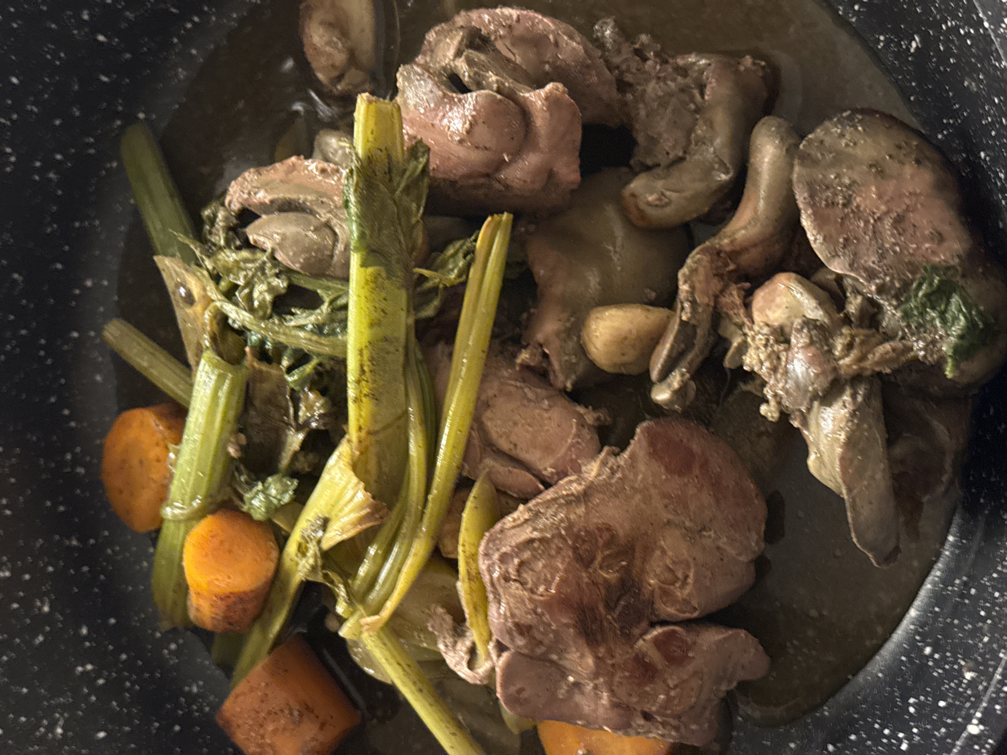 The image displays a close-up, overhead view of a cooked dish contained within a black, speckled cooking pan. The scene is situated in the city of Tel-Aviv, Israel. The primary subjects consist of numerous irregularly shaped pieces of dark, cooked meat, consistent with organ meat such as chicken livers, varying in color from reddish-brown to grayish-brown, with some showing textured surfaces. Interspersed with the meat are several bright orange, round-cut carrot pieces and multiple elongated, green stalks resembling celery, which appear partially cooked or wilted. Smaller, pale, ovoid components, potentially garlic cloves, are also visible among the ingredients. All elements are submerged in a shallow pool of dark, brownish liquid or sauce. The pan features a visible speckled pattern on its interior surface. No individuals or active actions are depicted, presenting a static view of the prepared meal.