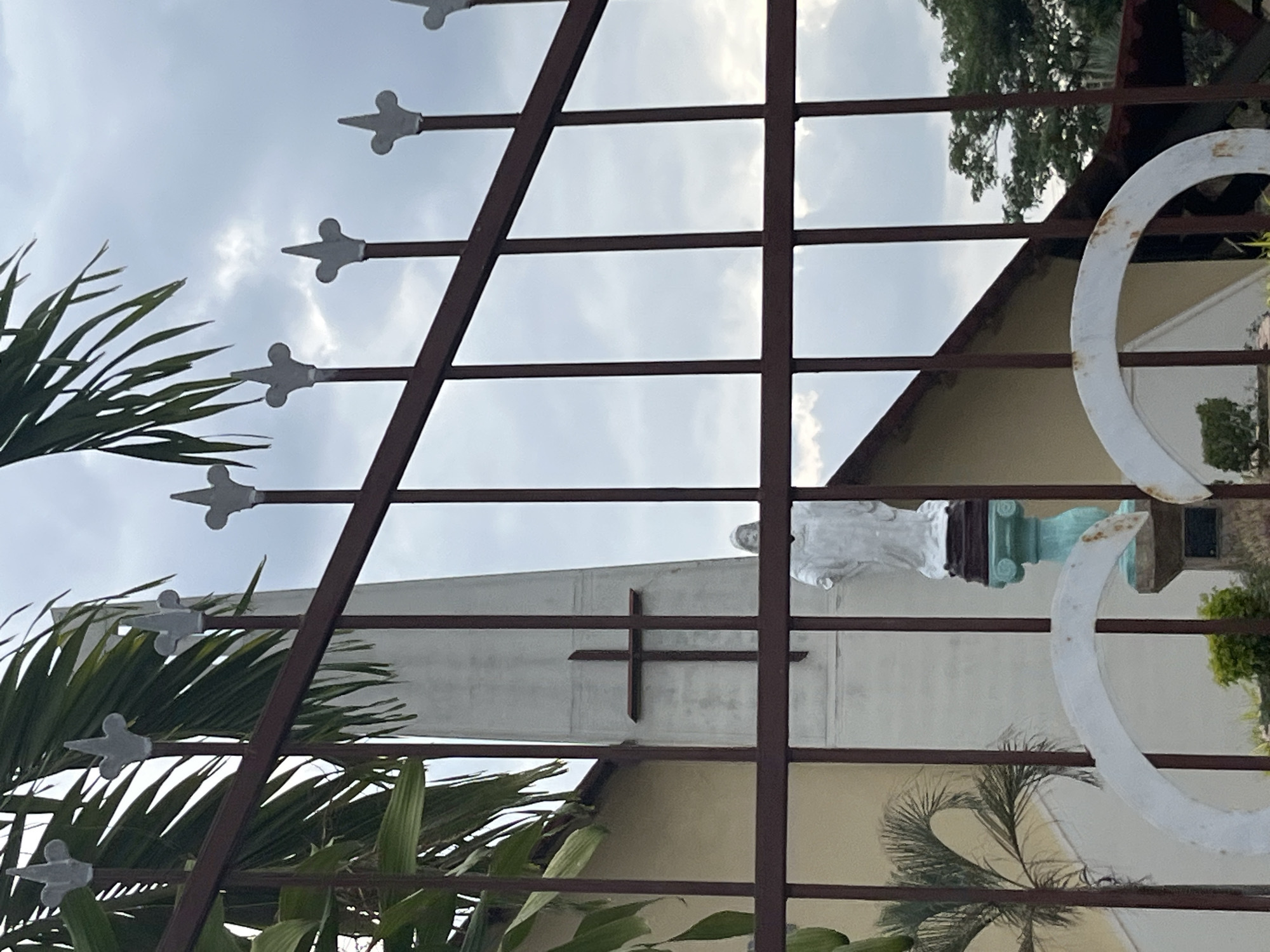 The image shows a partially obscured view of what appears to be a church or religious building, likely in Tinaquillo, Venezuela. The view is framed by a metal fence with decorative finials. Through the fence, a white building with a prominent cross on its facade is visible. A statue stands atop a pedestal, positioned against the building. Palm trees and other foliage are visible on either side of the structure. The sky is overcast. A white circular structure, possibly an architectural element, is seen on the right side.