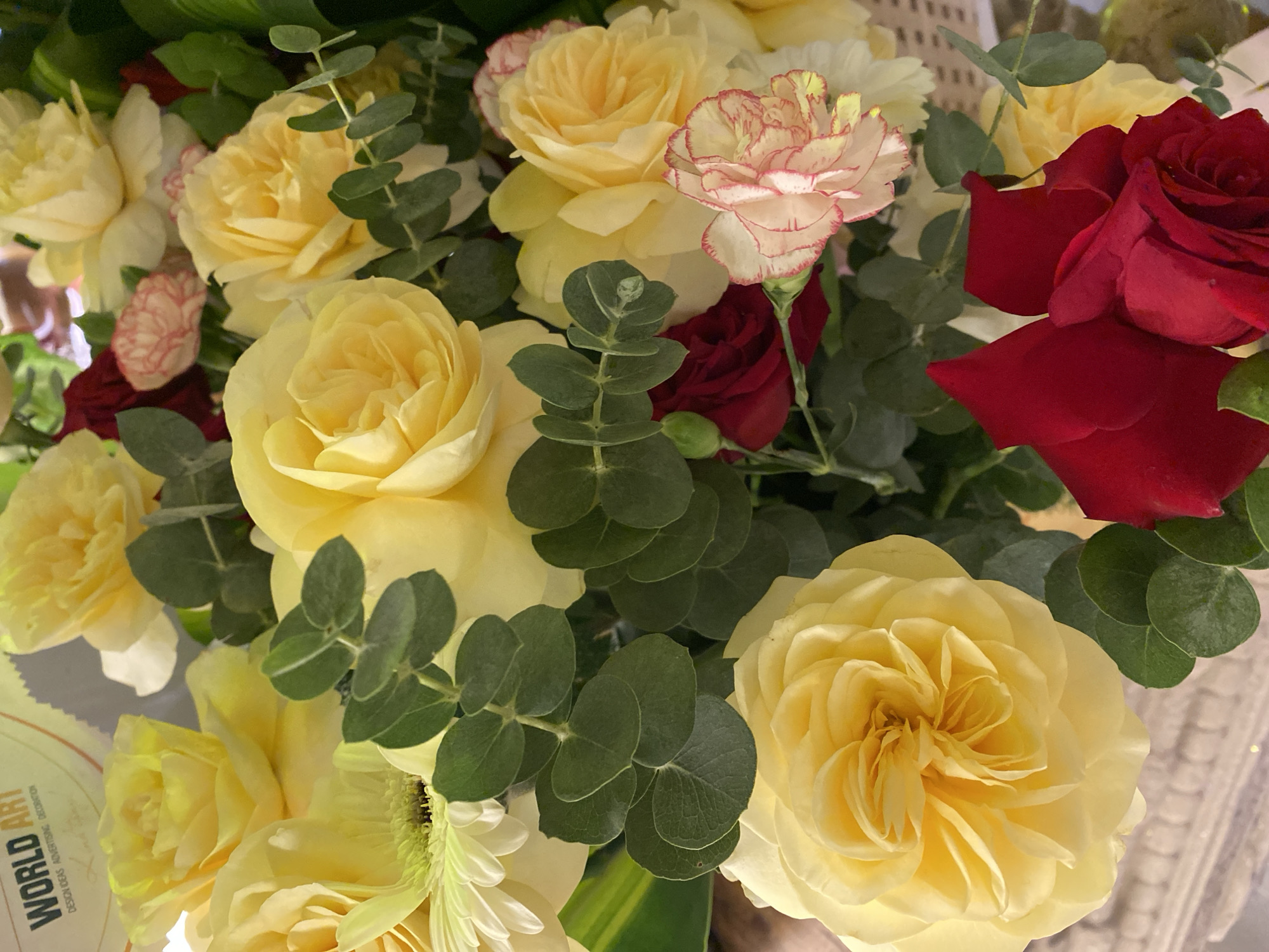 A close-up, overhead shot shows a bouquet of yellow roses, red roses, and pale pink carnations, interspersed with green eucalyptus leaves. In the upper left corner, a partially visible piece of paper or card displays the words "WORLD ART" and "DESIGN IDEAS ADVERTISING DECORATION". The background is blurred, but suggests an indoor setting. The lighting is bright, highlighting the textures and vibrant colors of the flowers. The scene is static, with no discernible action or human presence. This image could have been taken in Da Nang, Vietnam, where floral arrangements are common for both decorative and commercial purposes, such as in flower shops or event venues.