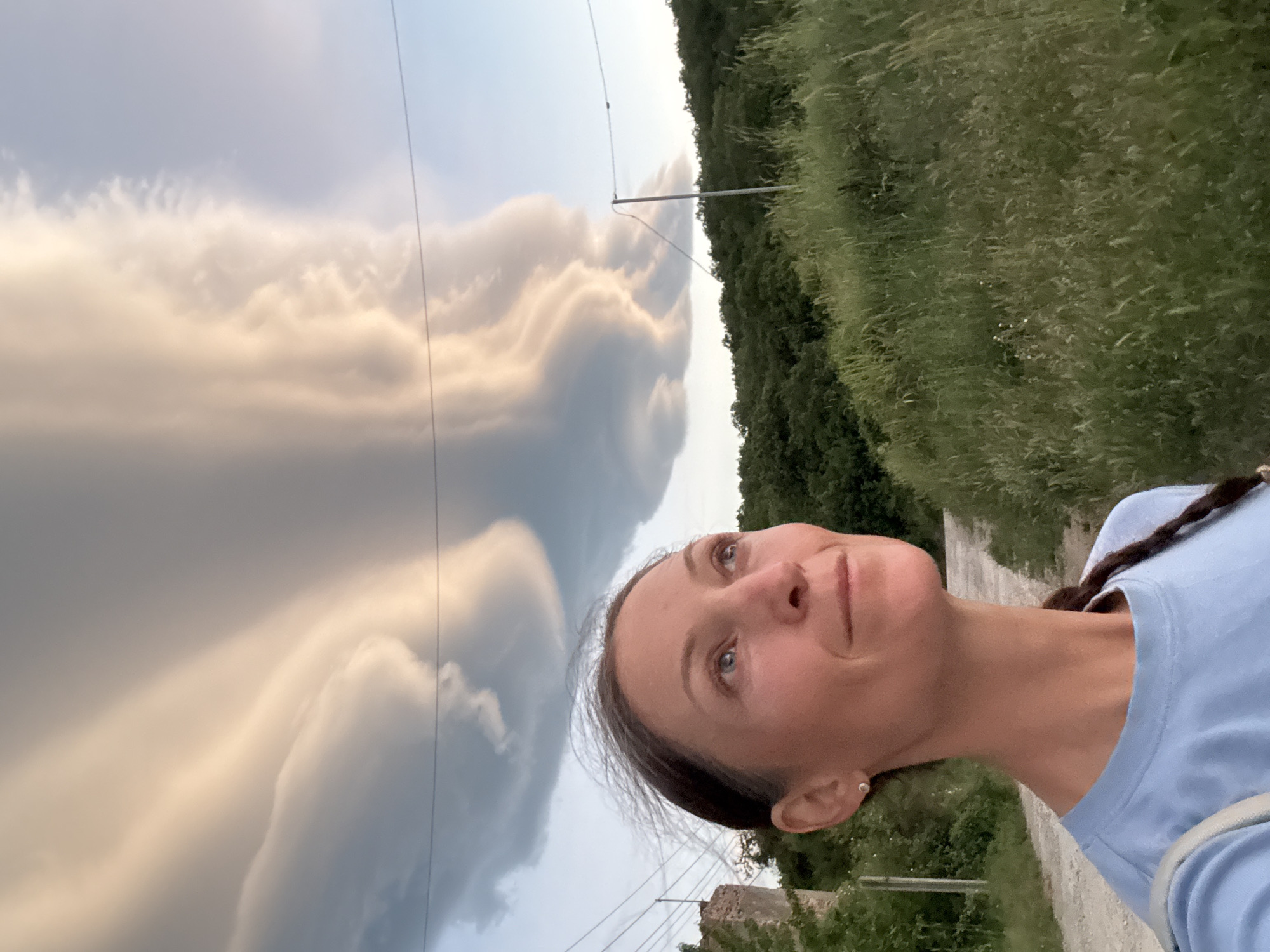 A medium shot depicts a woman outdoors, looking upwards and to the left. She has fair skin, blue eyes, and her hair is styled in a braid. She wears a light blue shirt.
The backdrop features a dramatic sky with shelf-like clouds. Electrical power lines are visible in the sky. A path or road borders the right side of the frame, flanked by greenery that includes grass and trees. The location is in the vicinity of the city of Chaika, Ukraine.