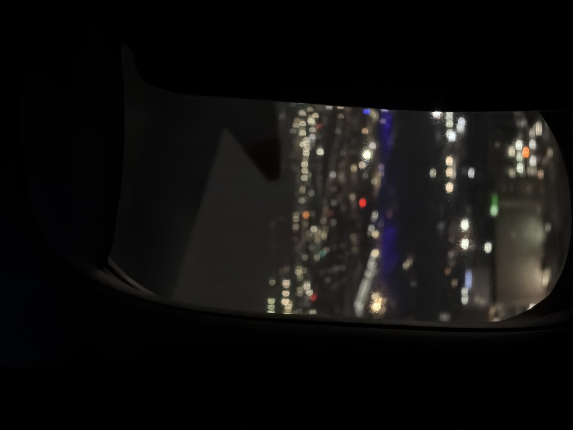 A blurred nighttime view of Los Angeles, USA, is seen through an airplane window. The city lights appear as indistinct bokeh, with vertical structures and illuminated buildings discernible through the window. Water droplets are present on the window. The interior of the airplane, surrounding the window, is dark.