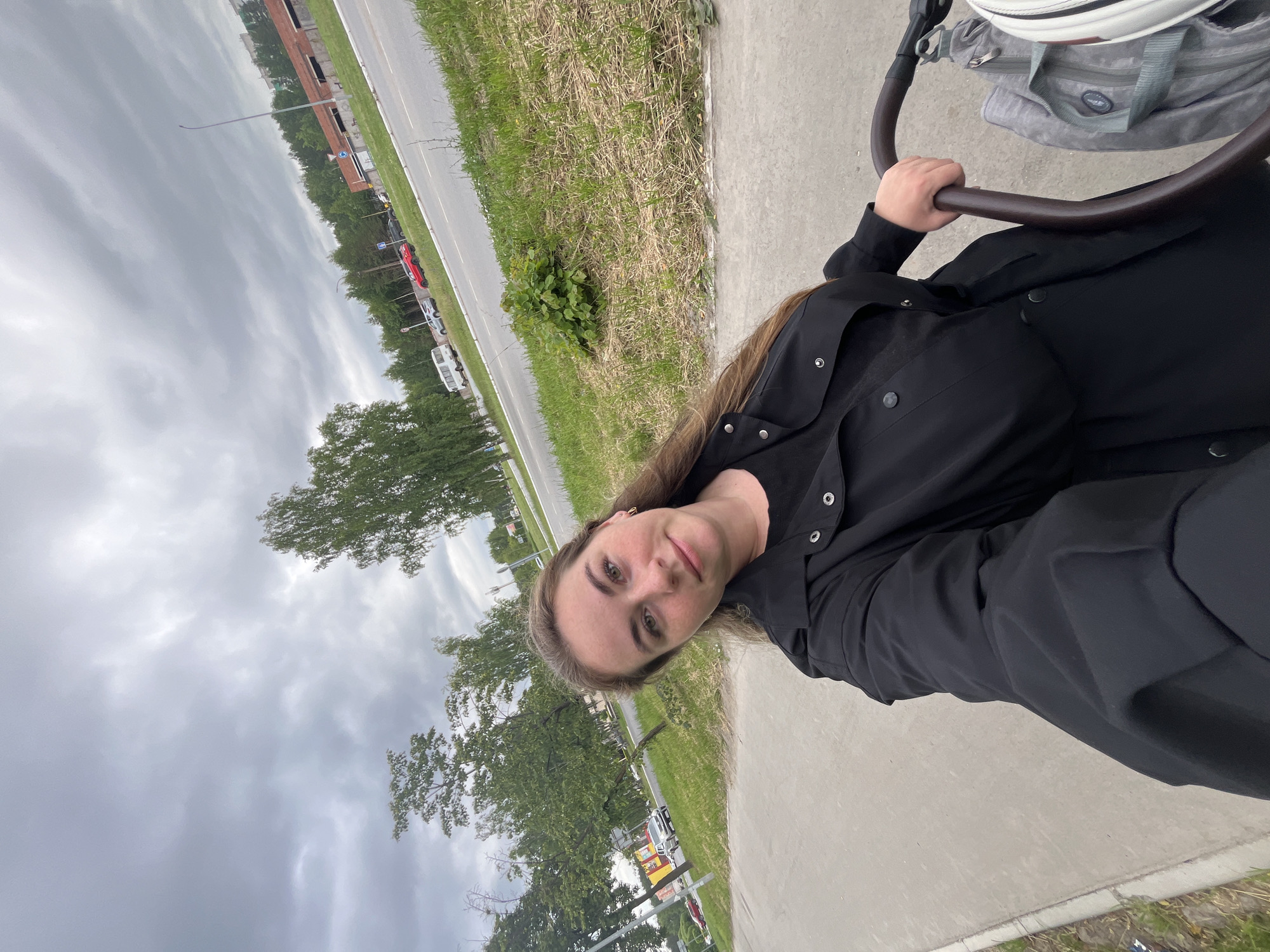 The media file shows a young woman taking a selfie outdoors. She is wearing a black outfit and has long brown hair. She is holding onto the handle of a stroller, suggesting she is with a baby or young child.
The scene appears to be along a paved path next to a grassy area and a road. Trees and some buildings are visible in the background, along with parked cars. The sky is overcast and cloudy, indicating it is likely daytime. The overall mood seems relaxed and casual.
There is no visible text in the image.