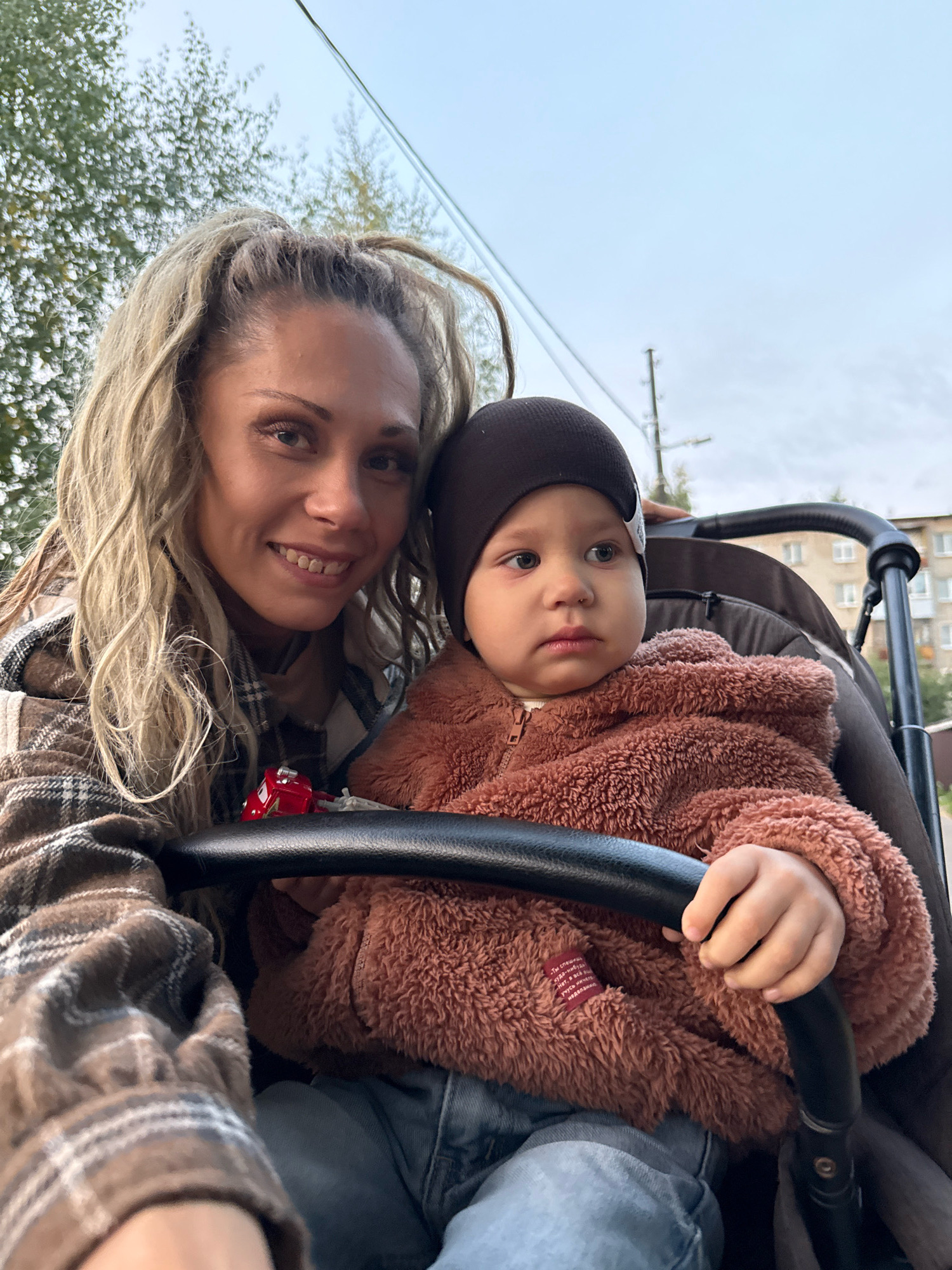 A medium shot shows a woman and a baby in a stroller, taken outdoors in Rusanovo, Russia. The woman is on the left, smiling and looking at the camera. She has blonde, braided hair and is wearing a brown and white plaid jacket. The baby is on the right, wearing a brown, fuzzy hooded jacket and a dark beanie. The baby is looking away from the camera. They are in a stroller with a black handle. In the background, there are trees and a building. The sky is overcast.