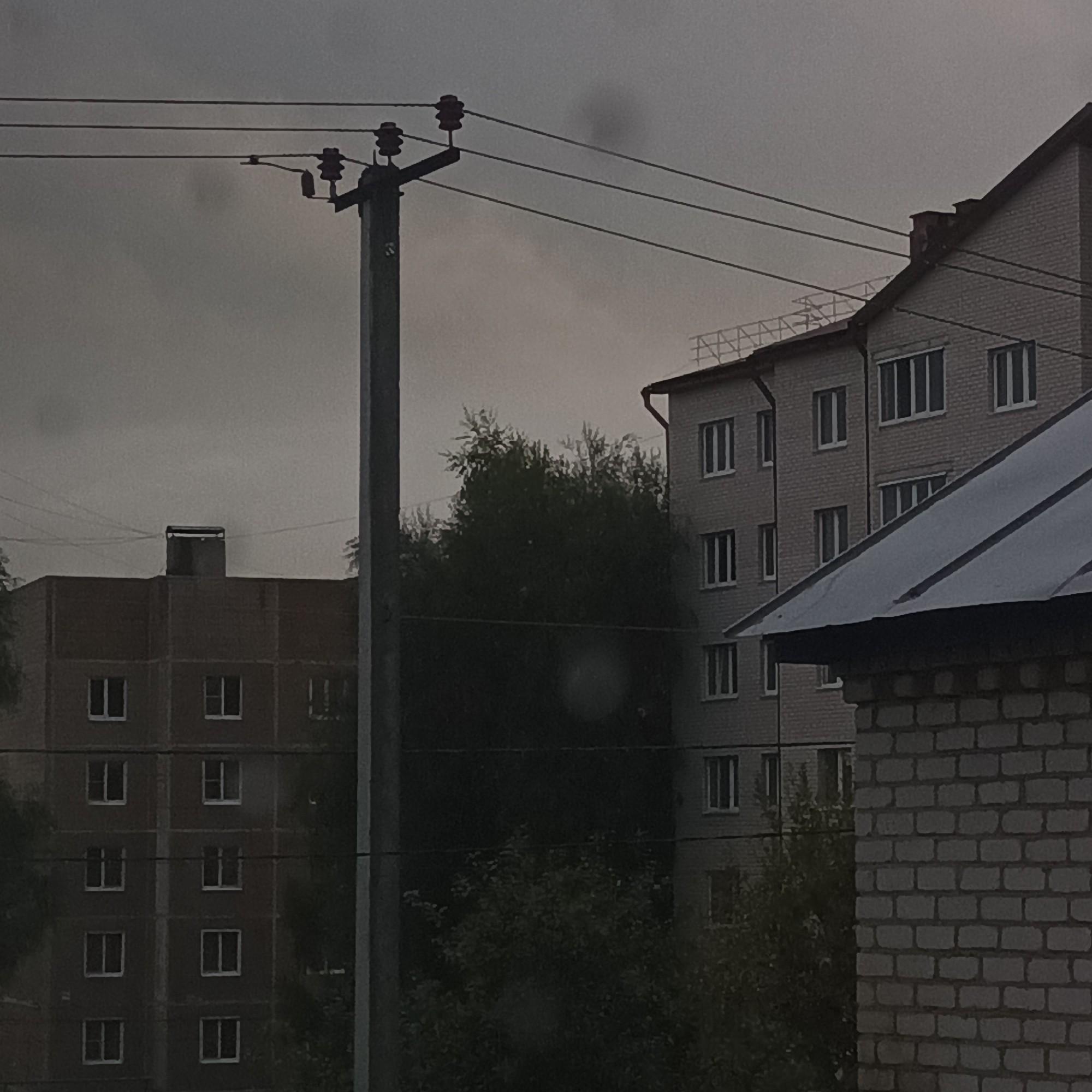 The image depicts an overcast sky above a street scene in Shumerlya, Russia. In the foreground, a utility pole with electrical wires runs vertically through the frame. To the left, a multi-story apartment building with a dark facade and numerous uniformly spaced windows is visible. To the right, another apartment building, constructed of light-colored brick, features multiple windows and a dark, sloped roof. A chimney rises from the roofline of this building. Dense, dark green foliage partially obscures the lower sections of both buildings and the space between them. The overall lighting suggests it is daytime, but the cloudy conditions create a somber atmosphere. There are no people or vehicles visible in the image.