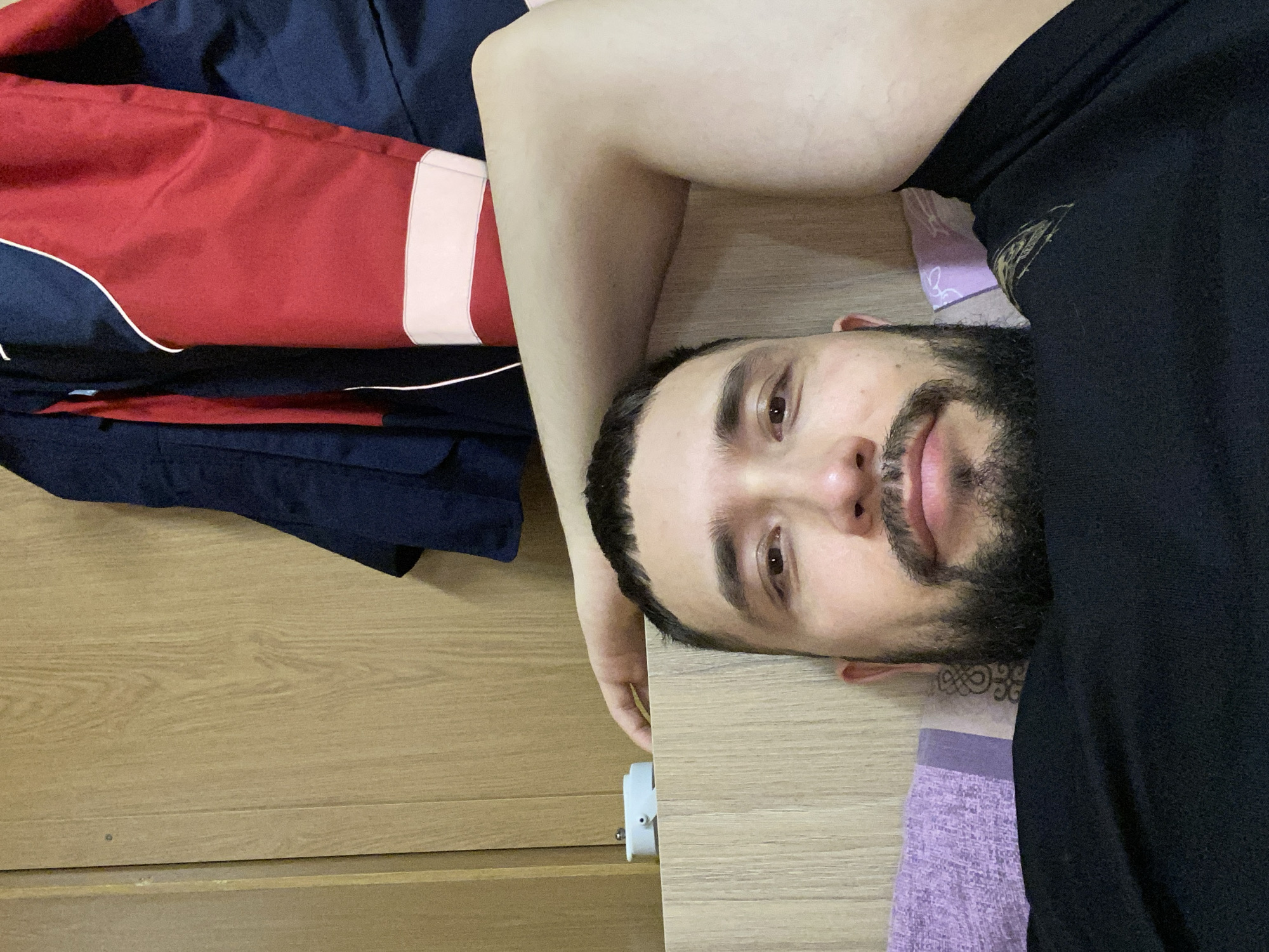 The image captures a man lying down and looking directly at the camera with a gentle, relaxed smile. He has short dark hair, dark eyebrows, and a neatly trimmed dark beard and mustache. A small, intricate tattoo, possibly an ornamental or Celtic knot design, is visible on his neck below his left ear. He is wearing a plain black t-shirt.
To his left, a folded garment, likely a sports jacket or tracksuit, in dark blue and red with white stripes and piping, is resting on a wooden surface. The man's head is resting on a light purple fabric with a subtle pattern, which appears to be a pillow or bedding. The primary background consists of light-colored wooden surfaces, suggesting an indoor setting such as a room with wooden furniture or flooring. A white cylindrical object, possibly an electrical plug or charger, is partially visible on the wooden surface near his left hand. The lighting is soft and even, consistent with an indoor environment during the day or in a well-lit interior. The overall mood is casual and friendly. There is no visible text in the image. This scene is set in Koskolovo, Russia.
