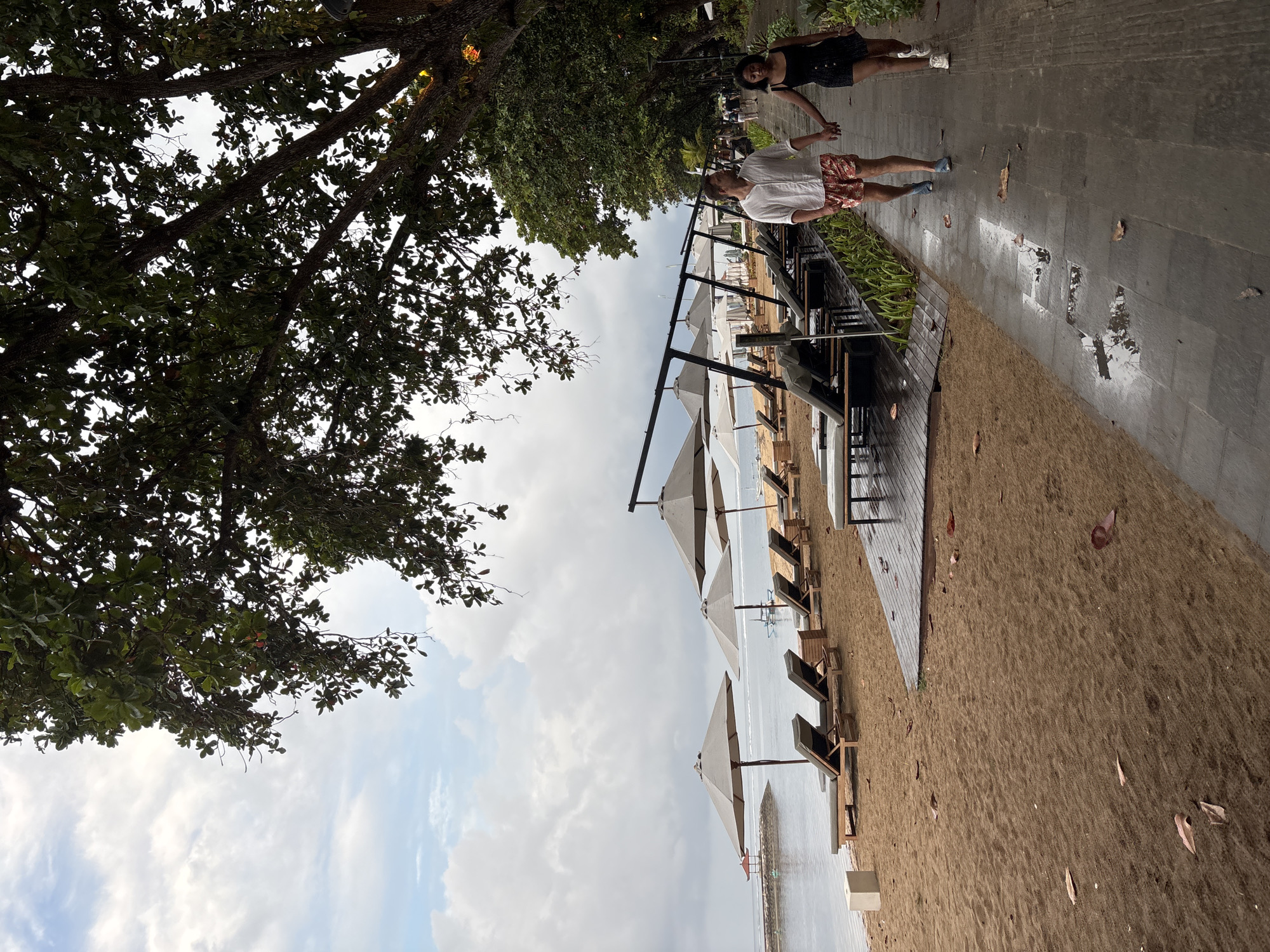 A couple, a man wearing a white shirt and patterned shorts, and a woman in a black dress, are walking hand-in-hand on a wet paved pathway. The pathway is lined with lush green plants on one side and a sandy beach with empty lounge chairs and umbrellas on the other. The beach is adjacent to a calm body of water, likely the ocean, under a cloudy sky. The scene appears to be a resort area in Denpasar, Indonesia, possibly on Kuta Beach or a similar coastal location, given the presence of beach amenities and tropical foliage. The overall impression is a serene, overcast day at a tropical beach resort.