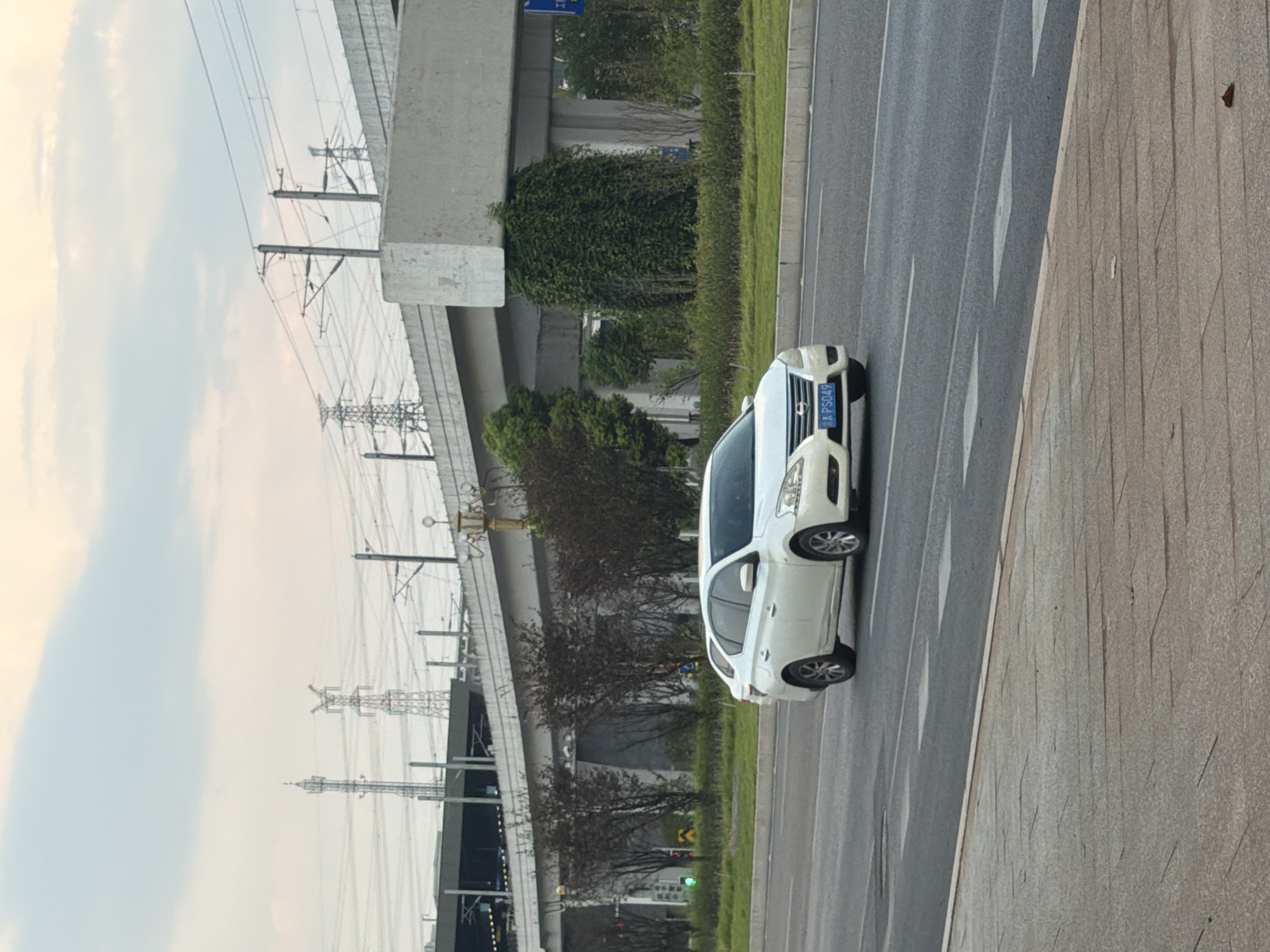 In Jiangjin, China, a white four-door sedan is driving on a paved road. The car has a blue license plate that reads "A PS049". The road has multiple lanes and is bordered by a sidewalk. In the background, an elevated structure, possibly a bridge or overpass, is visible. Power lines and transmission towers are also present in the distance. The sky is overcast with some clouds. The scene appears to be during the day.