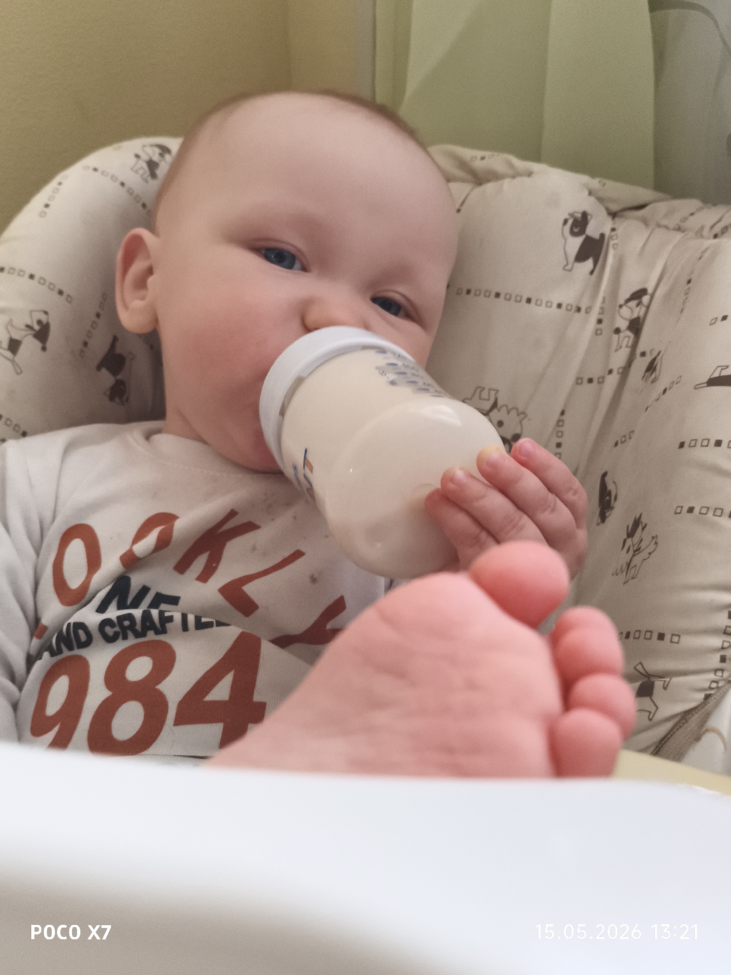 A baby is drinking from a bottle while seated in a high chair. The baby has light skin, blue eyes, and is wearing a white shirt with the words "BROOKLYN AND CRAFTL 1984" printed in red letters. The high chair has a patterned cushion with dog illustrations. The baby's foot is prominently displayed in the foreground, partially obscuring the view. In the bottom left corner, the text "POCO X7" is visible, and in the bottom right corner, the date "15.05.2026 13:21" is displayed. The background consists of a light yellow wall and a green curtain. The location is not discernible from the image.