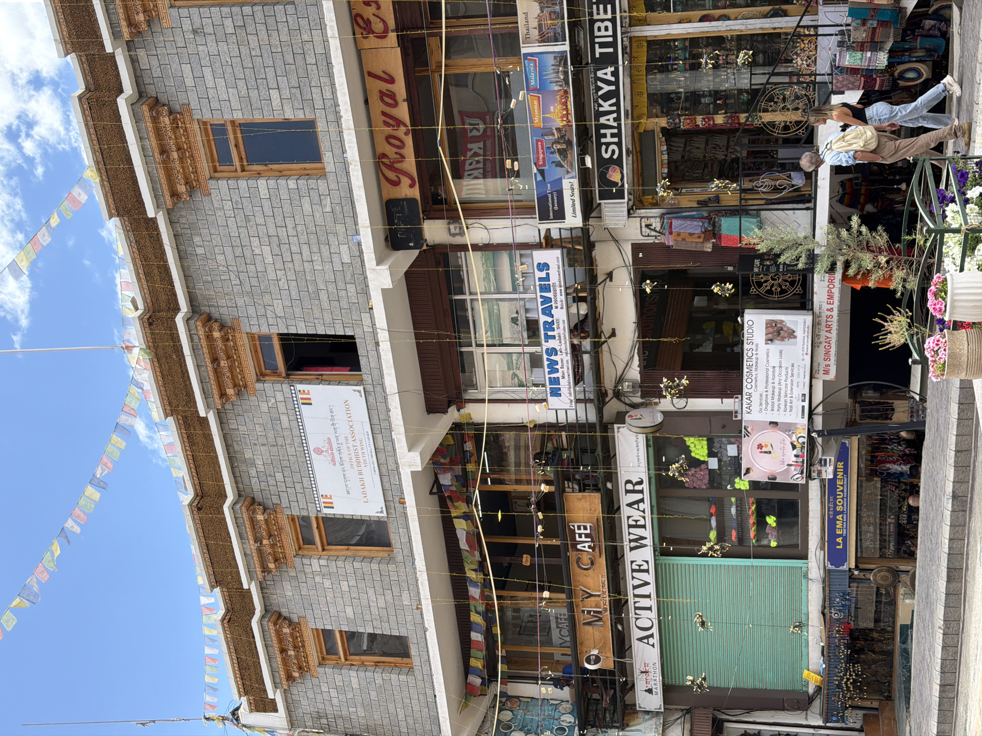 The image depicts a street scene in Leh, India, featuring the facades of multiple shops on a multi-story building. Prayer flags are strung across the upper levels of the building and across the street. The building's architecture includes stone brickwork, wooden window frames, and decorative elements.
On the ground floor and extending to the first floor, several businesses are visible. From left to right, there's a shop with a green shuttered entrance labeled "ACTIVE WEAR". To its right, a shop called "LA EMA SOUVENIR TIBETAN AND LADAKH HANDICRAFT" displays various handcrafted items. Further to the right, a shop labeled "KAKAR COSMETICS STUDIO" offers beauty services. Next to that, a shop called "SHAKYA TIBET" displays textiles and handicrafts.
Above these ground-floor establishments, on the first floor, are more shops. One is "MY CAFE," with outdoor seating visible. Adjacent to it is "NEWS TRAVELS," which has a display of travel destinations. To the right of that, a shop with "Royal Es" visible on its signage appears to be a business, possibly a restaurant or store.
In the foreground, two individuals, a man and a woman, are walking away from the camera on a paved sidewalk. The man is wearing a blue shirt, beige pants, and a backpack, while the woman is wearing a black tank top and light blue jeans. Flower pots with pink and white flowers are situated on the sidewalk. The environment is characteristic of a bustling market street in Leh, with shops selling local crafts, clothing, and travel services, under a clear blue sky with scattered clouds.