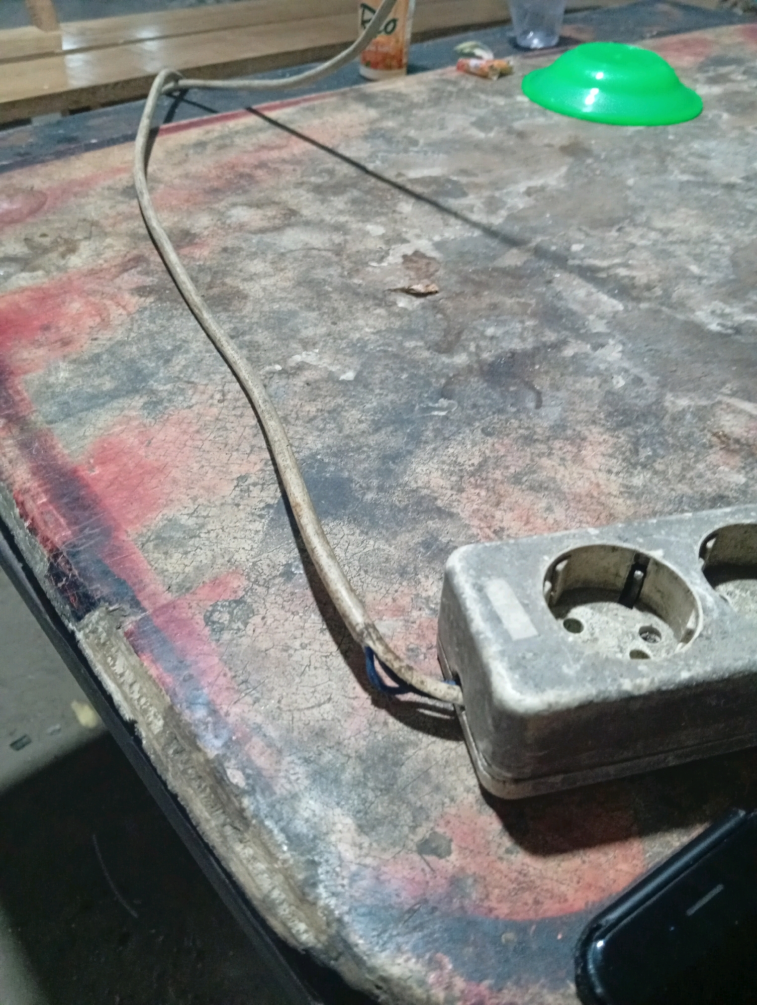 The image depicts a close-up, high-angle shot of a worn and dirty table surface. The table appears to be made of a dark material with visible red and pink stains, possibly paint or rust, and has a rough texture. A thin, grey electrical cord extends diagonally across the table from the lower left towards the upper right. A grey, multi-socket electrical extension cord is positioned on the right side of the table, with its outlets facing upwards. The extension cord shows signs of dust and age. In the background, out of focus, are wooden planks, a clear plastic cup, and a bright green cap or lid. A small, dark object, possibly a smartphone, is partially visible in the lower right corner of the image, with its screen facing downwards. The overall scene suggests a cluttered or utilitarian workspace. The location is identified as Kubang Jaya, Indonesia, though no specific landmarks are visible in the image to confirm this.