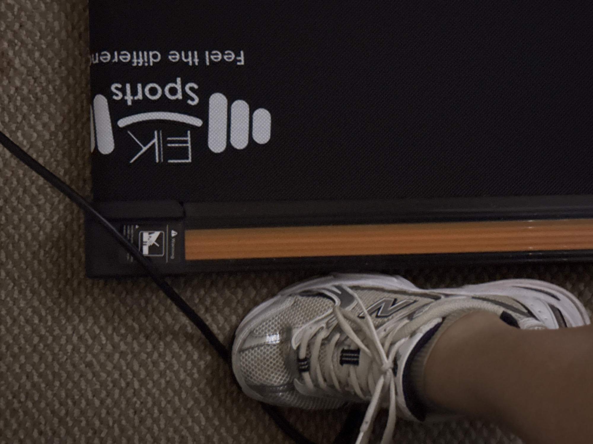 The image captures a close-up, angled view of a person's foot, wearing a white and grey New Balance sneaker, positioned on a textured, light brown carpet. The foot is situated next to a black fitness machine, likely a walking pad or compact treadmill, suggesting preparation for or engagement in exercise. An electrical cable extends from the lower-left corner of the frame, running across the carpet and along the side of the machine.
The fitness machine features a black surface with branding in white. Clearly visible is the text "EK Sports" accompanied by an emblem resembling a stylized barbell or dumbbell. Below this, partially cropped, are the words "Feel the differen", which likely completes the phrase "Feel the difference". A small warning label is present on the side of the machine, displaying an icon that depicts a hand being pulled into a mechanism, along with the text "Warning". An orange-brown strip runs along the edge of the machine's base.
The scene is indoors, characterized by the carpeted floor. The lighting appears to be ambient, offering no specific cues about the time of day. The location, Great Yarmouth, United Kingdom, is implied by the context provided, suggesting a common home exercise setup. The image focuses on the immediate interaction between the user and the exercise equipment.