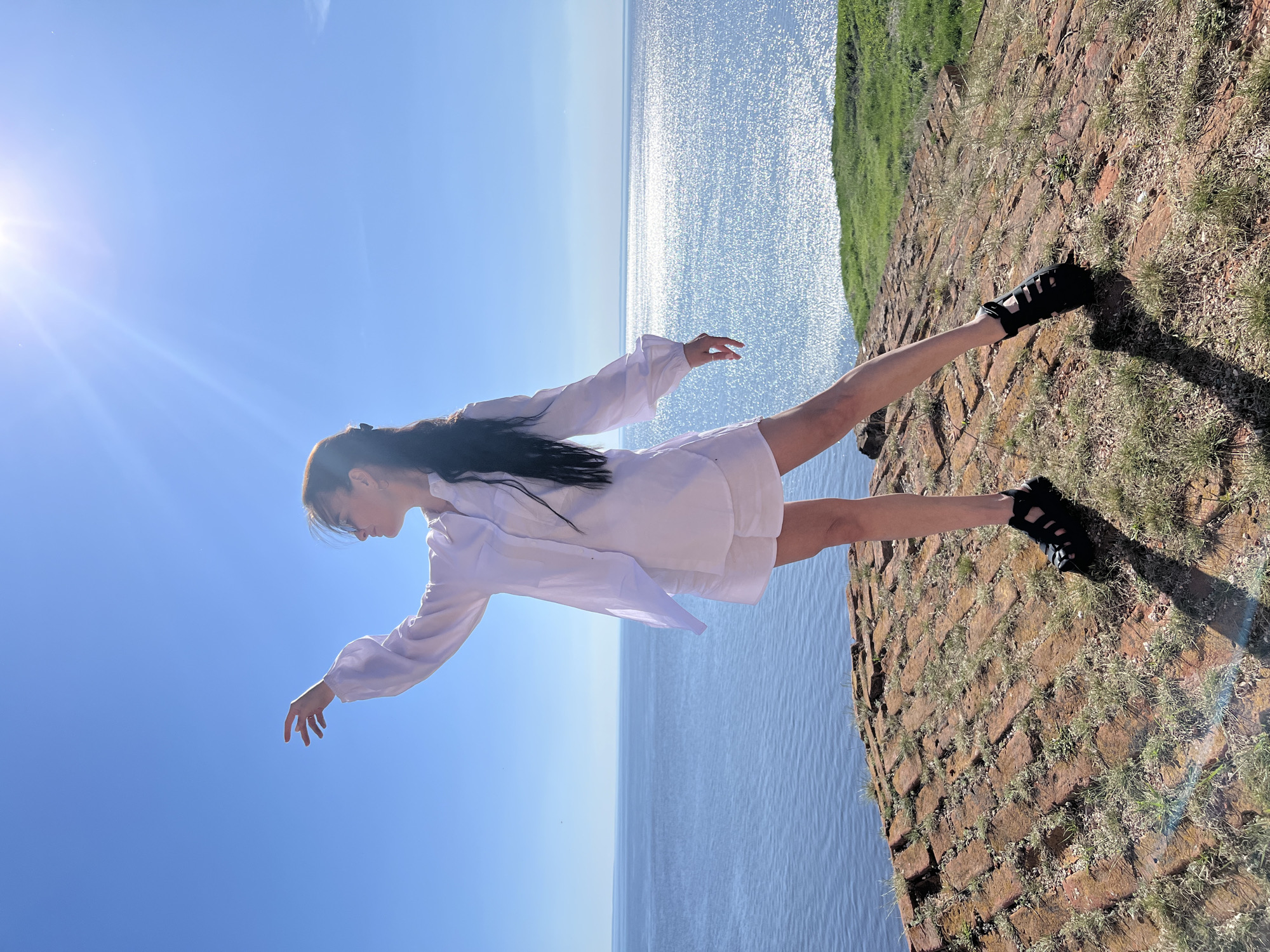 A vertical outdoor shot features a woman with dark hair wearing a white dress and black sandals. She stands on a brick and grass surface, seemingly near a body of water that stretches into the distance under a clear blue sky. The shot appears to be taken in Шуран, Russia, based on location. The sun's glare is visible in the upper left of the frame.