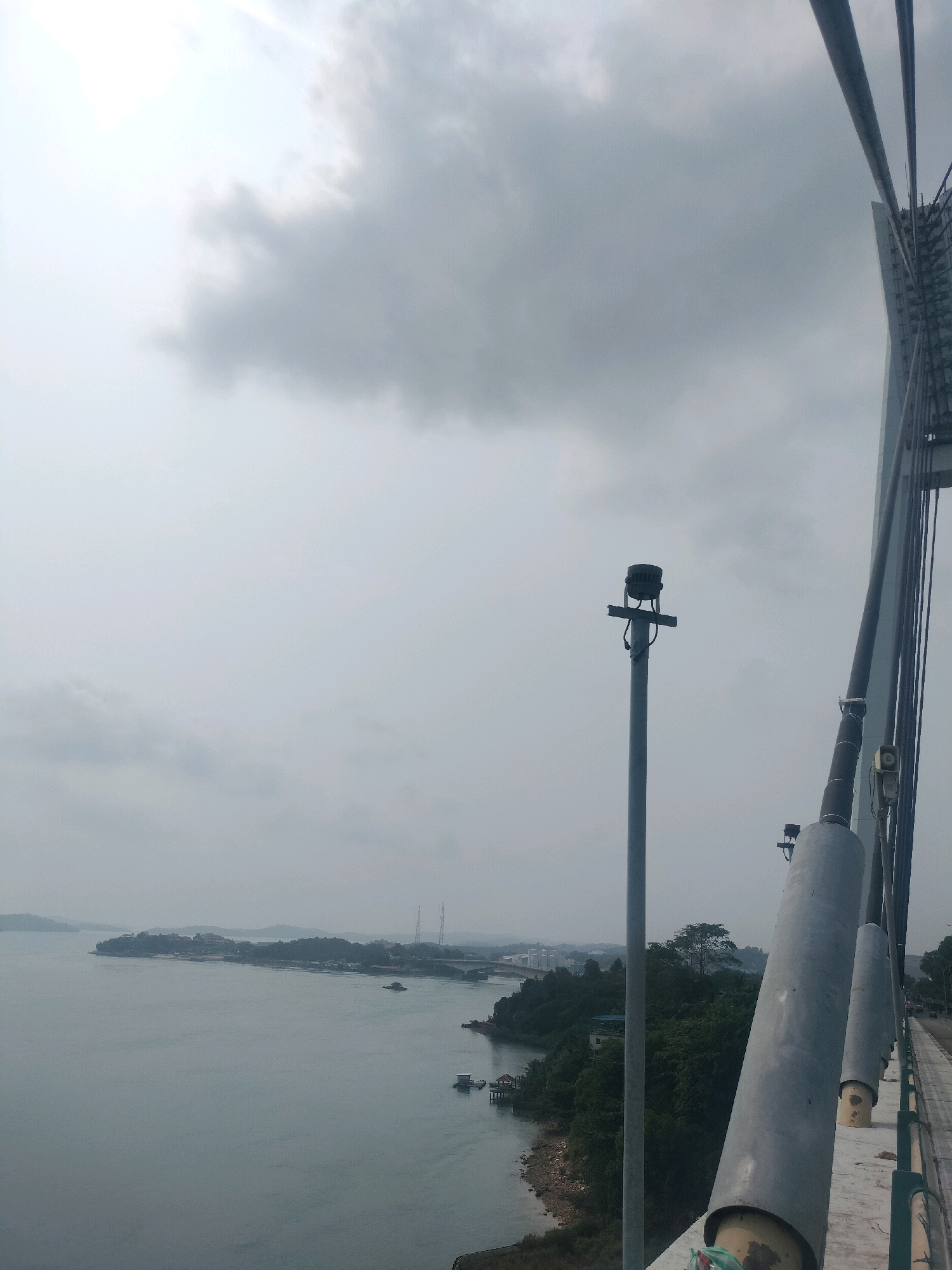 The image shows a view from a bridge overlooking a body of water with islands and a coastline in the distance. The sky is overcast with gray clouds. On the bridge, there are structural elements, including large cables and support pillars. A tall pole with a light fixture is visible on the left side of the frame. In the distance, there is a shoreline with trees, buildings, and what appear to be more bridges or elevated structures. A few boats are present on the water, and a small structure resembling a floating house or platform is visible closer to the shore. The overall impression is of an industrial or transportation infrastructure area within a coastal or island environment. This scene is consistent with the landscape of Batam, Indonesia, which is known for its bridges connecting its many islands.