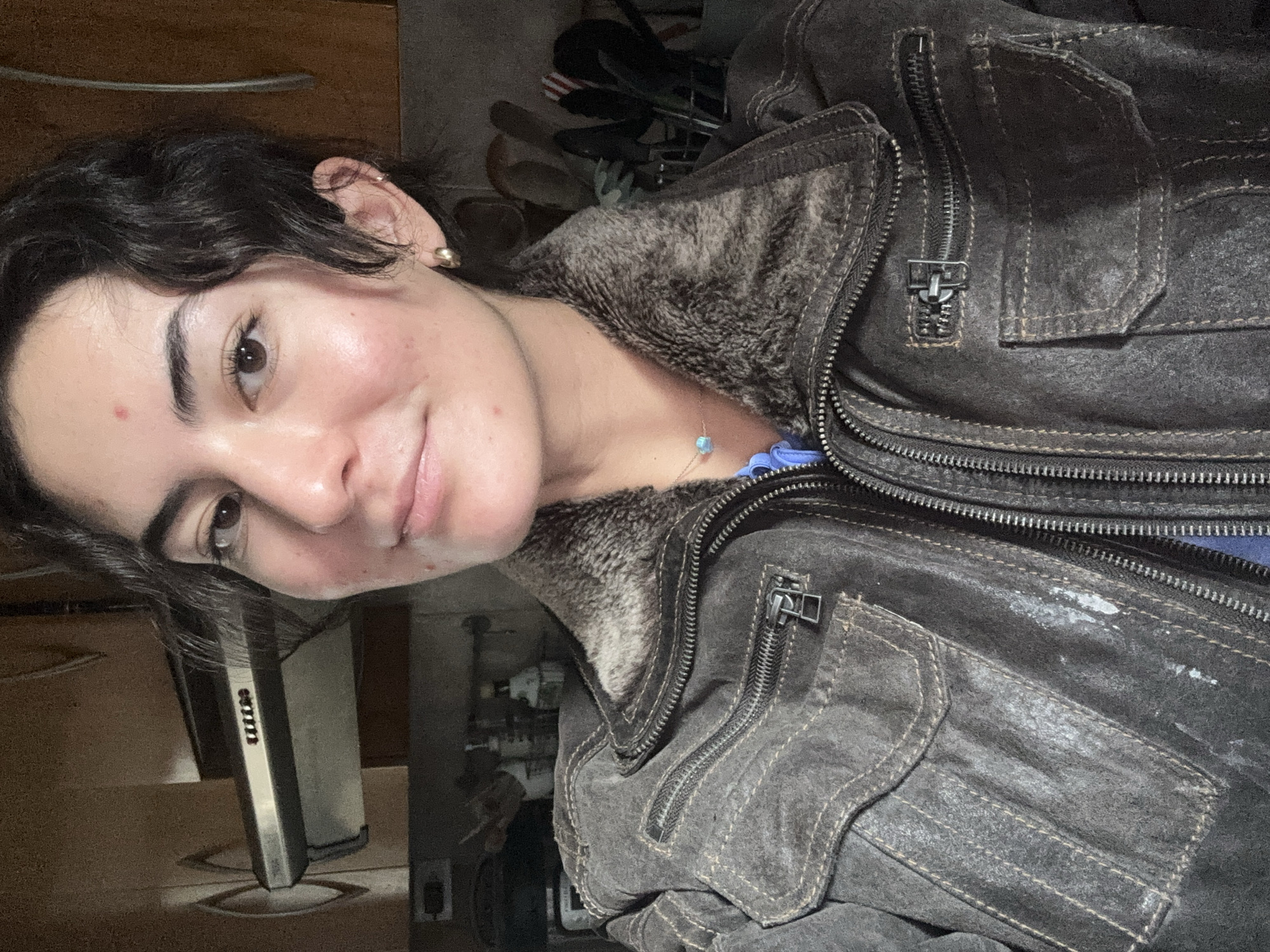 A selfie portrait of a fair-skinned person with dark brown hair and eyes, wearing a dark brown leather jacket with a fur-like collar, taken indoors. The person has a small gold earring in their left ear and is wearing a blue top under the jacket. A delicate necklace with a small blue pendant is visible. In the background are various indistinct objects including what appears to be a shoe rack, a metal object, and part of a door or cabinet. The image shows no elements that provide information regarding the location being the city of Puebla, Mexico.