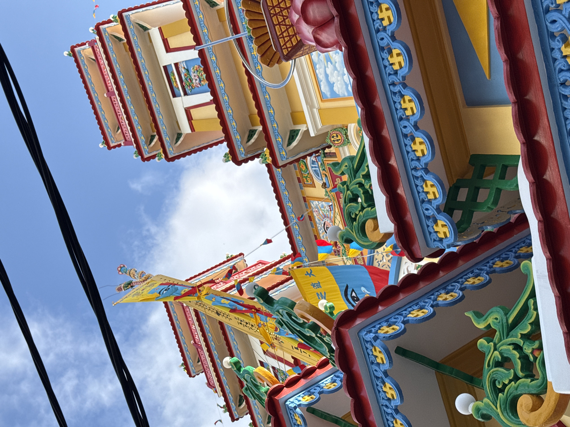 The image shows a portion of a brightly colored Buddhist temple in Nha Trang, Vietnam, under a blue sky with scattered clouds. The architecture is ornate, featuring multiple tiered roofs with red trim and decorative blue and yellow patterns, including swastika symbols. Vibrant yellow flags with religious imagery, such as a large blue eye and Chinese characters, are prominently displayed. Green dragon-like sculptures with white spherical eyes adorn the roof edges. The temple's walls are painted yellow and white, with some areas featuring painted murals of clouds and possibly landscapes. Black electrical wires are visible crossing the upper portion of the frame, suggesting the temple is in an urban or semi-urban setting. No people are visible in the image.