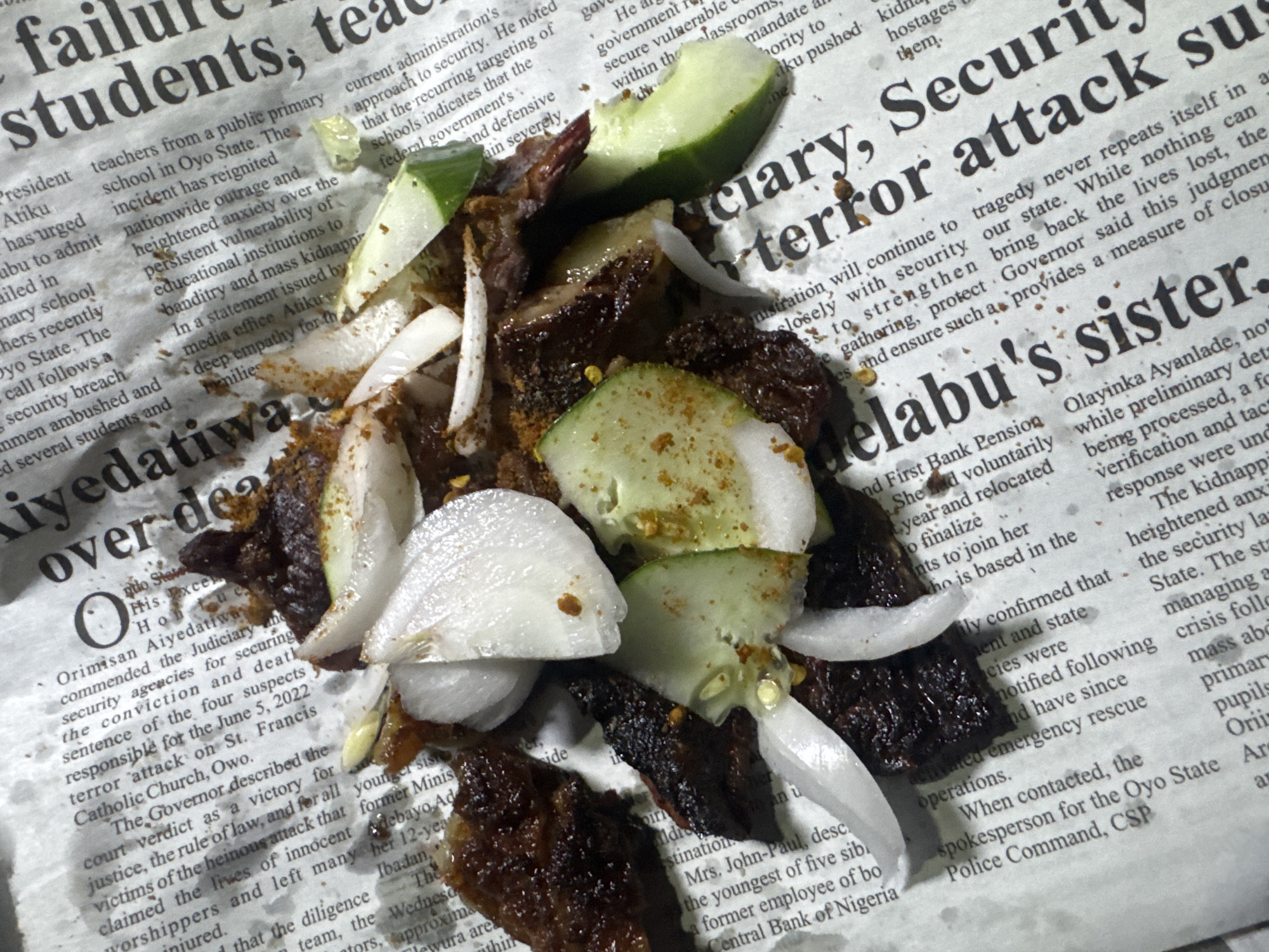 The image shows a close-up overhead view of food, likely a salad or appetizer, scattered on a crumpled newspaper. The food consists of dark meat chunks, sliced white onions, and cucumber slices, sprinkled with spices. The newspaper is partially visible, with text and headlines in English. One prominent headline reads "Students, teach...", suggesting it is a news article. The text indicates the location of a news report is in Oyo State, Nigeria, but there is no indication of Abuja.