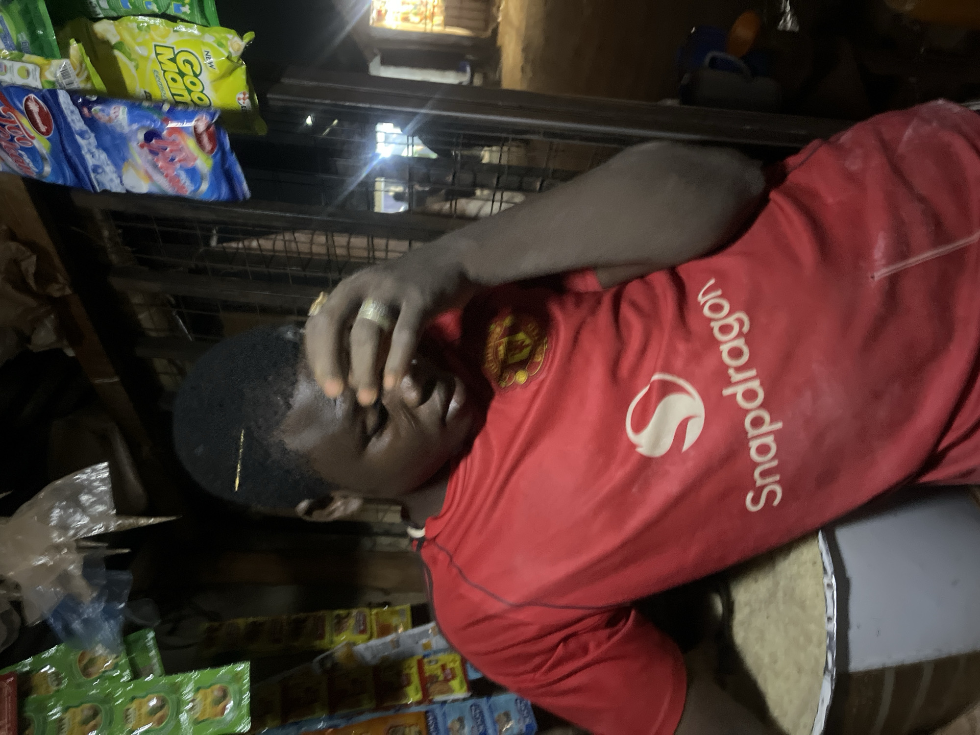 An individual wearing a red Manchester United jersey with white "Snapdragon" branding on the sleeve is resting their head on their hand, possibly in distress. They are seated next to what appears to be a gray bucket or container. The scene is in what looks like a small store or shop, with shelves stocked with various snacks and household products. The location is estimated in Abuja, Nigeria. The overall lighting is dim, suggesting it is indoors or at night.