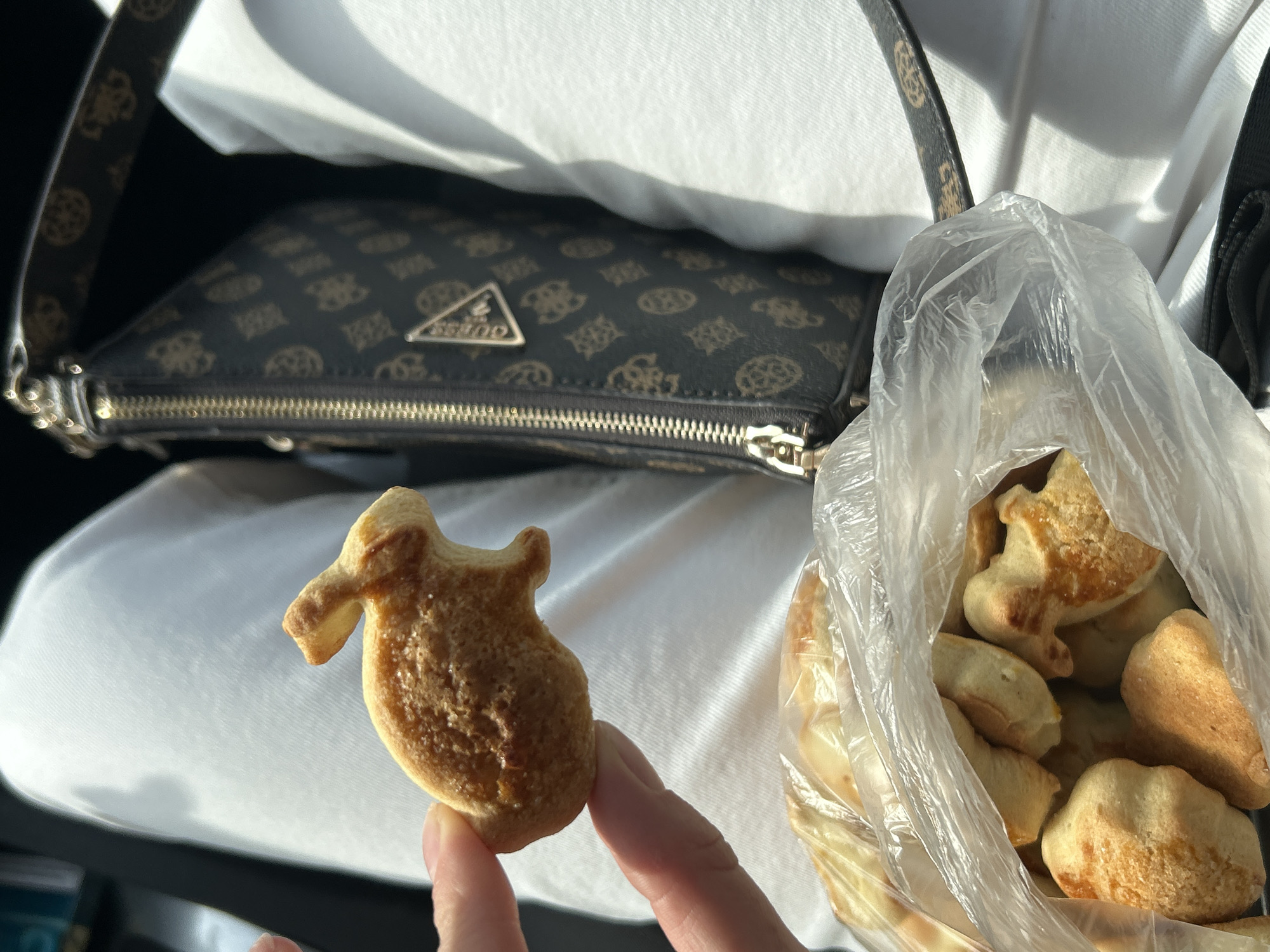 The image shows a hand holding a small, oddly shaped cookie with a textured surface, in front of a clear plastic bag filled with similar cookies. Behind the hand and bag is a patterned purse, presumably made of synthetic material, resting on a white garment. The context suggests the setting is likely indoors, possibly inside a vehicle. There is no confirmation the image was taken in Церово, Bulgaria.