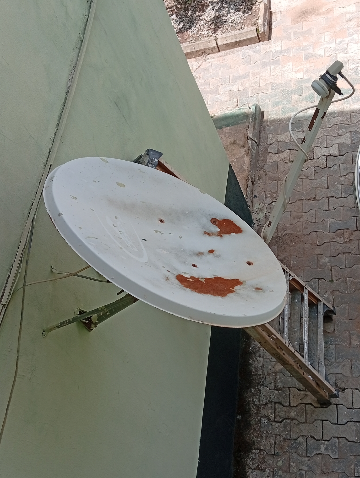 The image depicts a satellite dish attached to the side of a pale green building. The satellite dish is white with patches of reddish-brown staining. The mounting bracket is green and appears weathered. A ladder leans against the building near the satellite dish. The building is adjacent to a paved area composed of interlocking concrete blocks. The scene is in Dutsen Alhaji, Nigeria.