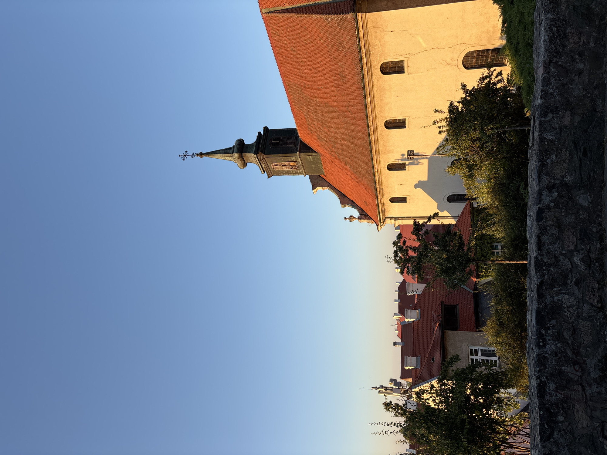The image captures a section of Petrovaradin, Serbia, with a focus on historic architecture bathed in the warm glow of late afternoon sunlight. A pale yellow building, possibly a church, dominates the right side of the frame, its terracotta tiled roof catching the light. The building features arched windows and a steepled tower topped with a cross.
In the background, a cluster of other buildings with red tile roofs are visible, interspersed with lush green trees. A rough stone wall runs along the bottom foreground, adding a textural contrast to the smoother surfaces of the buildings and the clear blue sky above. The overall impression is one of peaceful, historic urban landscape at sunset. There are no people or active events depicted in the image.