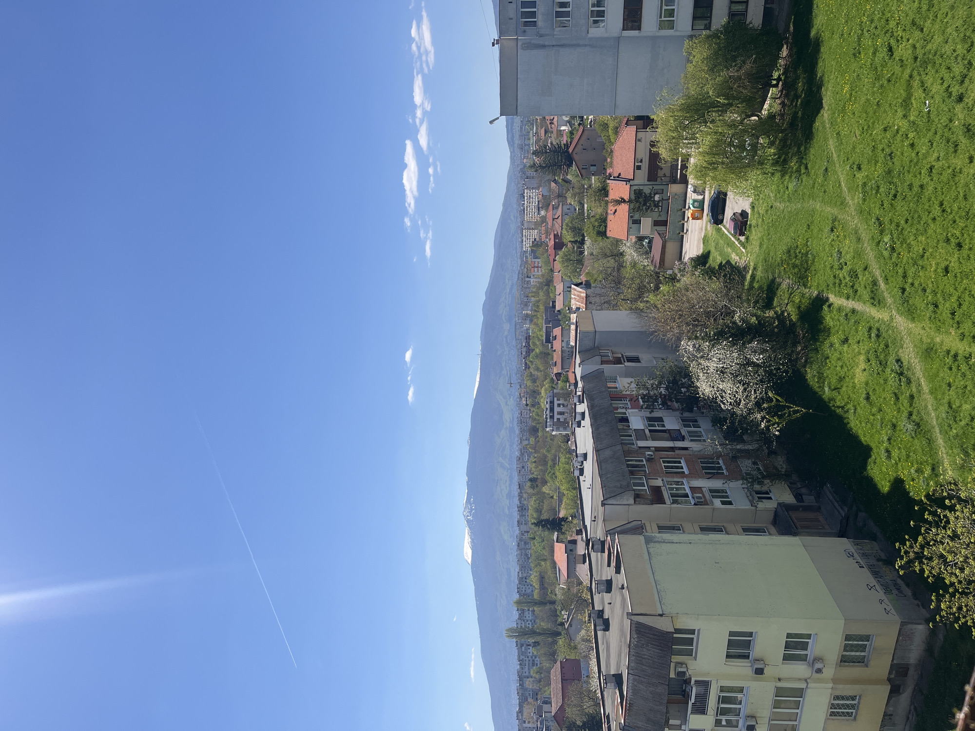 The image is a high-angle shot of an urban landscape, likely taken from an elevated position such as an upper-story building or a high point. The location appears to be in Sofia, Bulgaria, identifiable by the distinct mountain range in the background, resembling the Vitosha Mountain. The city sprawls between the viewer and the mountain, displaying a mix of apartment buildings, residential houses with red-tiled roofs, and green spaces.
In the foreground, the roofs of buildings, including one with graffiti on its side, are visible. A grassy area with a worn path running through it is located beside buildings. Several cars are parked nearby. Above, a clear blue sky occupies a significant portion of the frame, marked by a contrail from a high-flying aircraft.
The overall scene presents a typical urban setting in Sofia, where residential areas are nestled near natural landscapes.