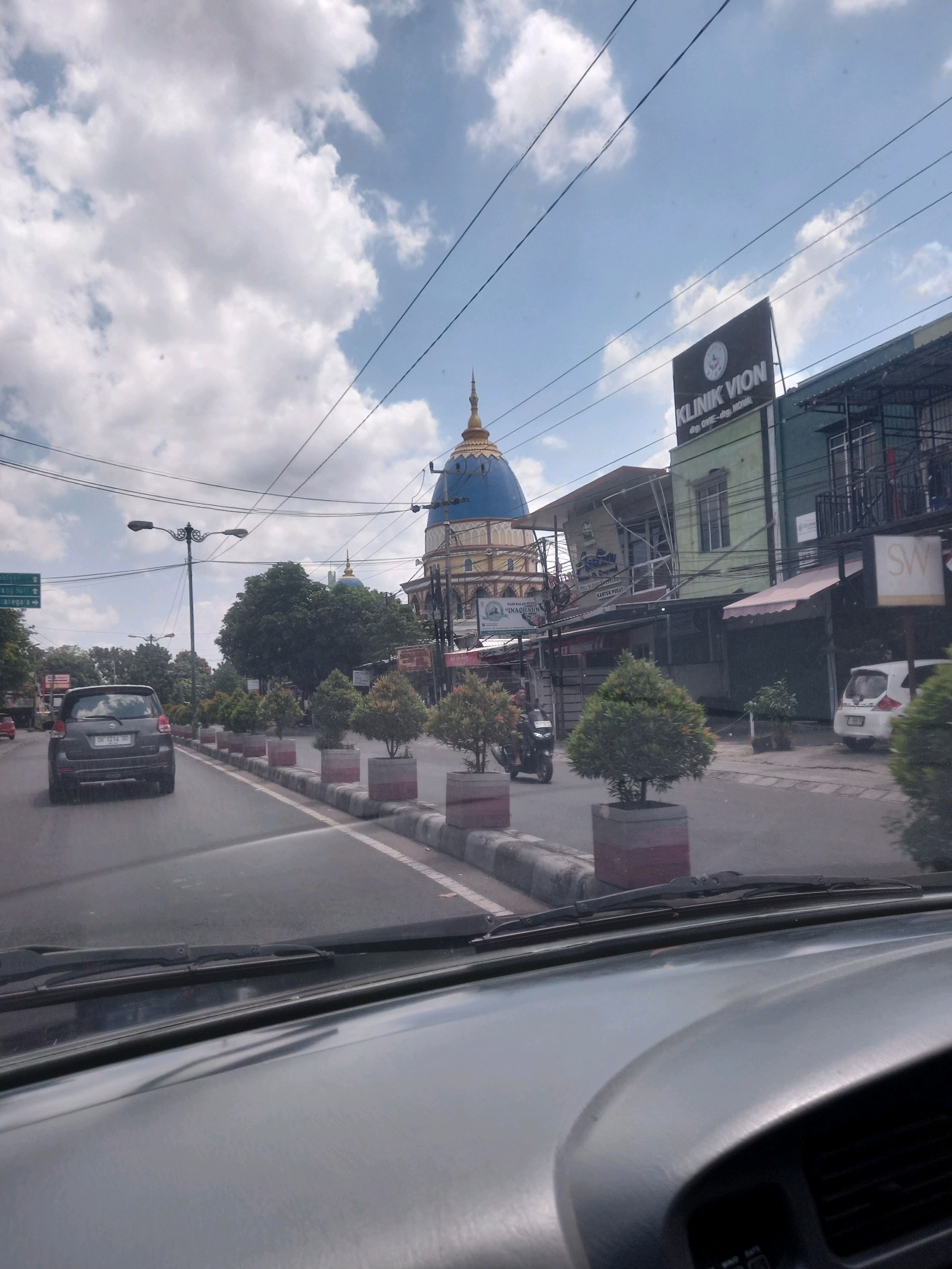 The image shows a street scene in Mataram, Indonesia, likely on a bright, cloudy day. In the foreground, the dashboard of a vehicle is visible, with a windshield wiper partially obscuring the view of the road ahead. A dark-colored hatchback is driving in the left lane. The road is lined with potted plants and has a pedestrian walkway.
Dominating the background is a striking blue domed building with gold accents, likely a mosque or a significant religious structure, possibly the Grand Mosque of West Nusa Tenggara. To the right of this building are several businesses, including one with a prominent green sign that reads "KLINIK VION". The sky is filled with dramatic white clouds against a blue background, with power lines crisscrossing the scene. The overall impression is a bustling urban environment with distinctive architectural features.