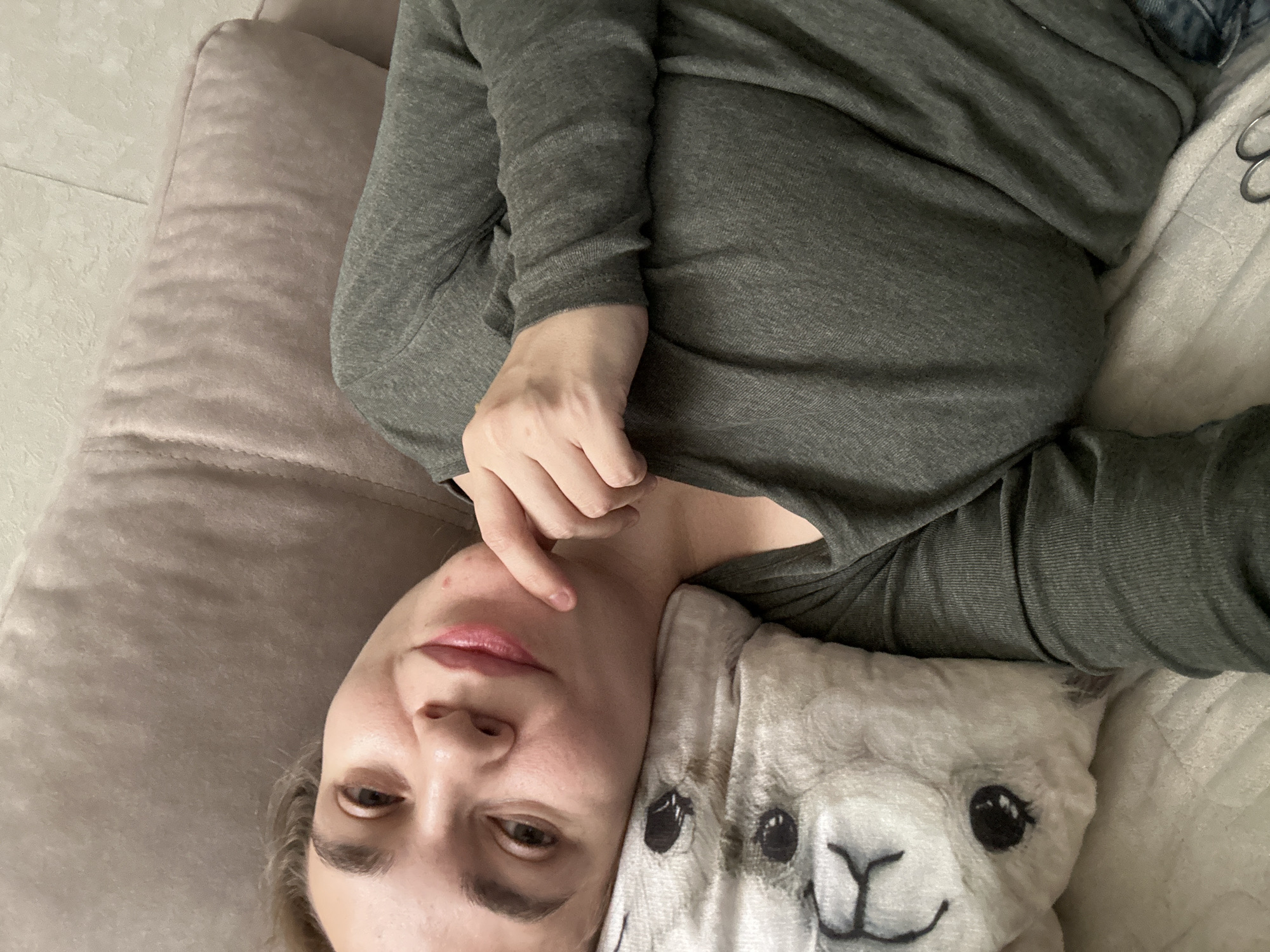 A person lies on a light beige cushion with an alpaca-printed pillow to the right. The person wears a long-sleeved, gray-green shirt. The person's face is visible, with their right hand touching their cheek. The background appears to be a light-colored floor. The exact location of the image is unknown. It is not possible to determine if this image was taken in Uva, Russia, or anywhere else.