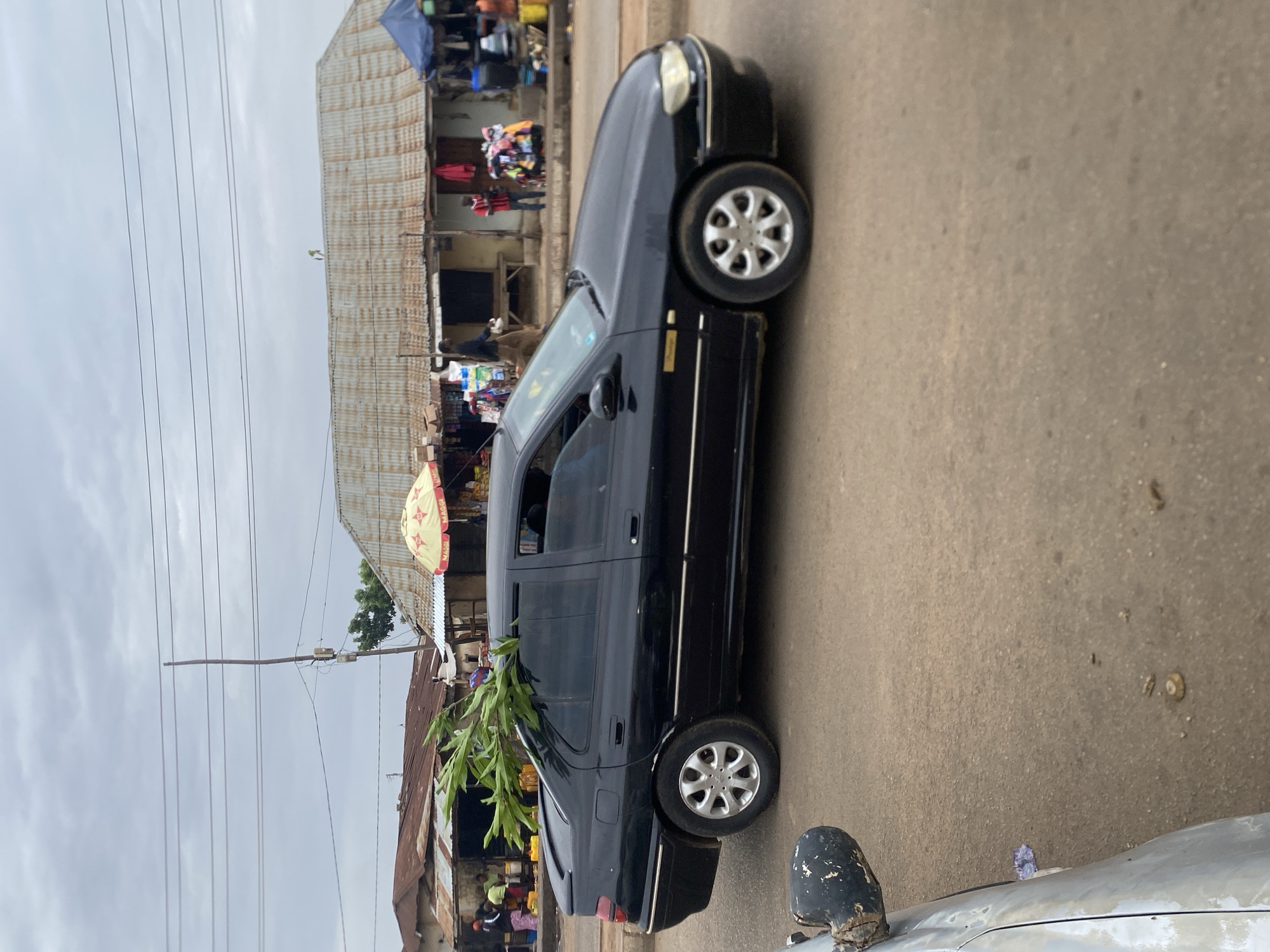 The image shows a black sedan parked on a paved road in Bukuru, Nigeria. The car has foliage sticking out from its side windows. In the background, there are structures with corrugated metal roofs and people engaged in market activities. Utility poles with visible wires are also present. The sky is overcast.