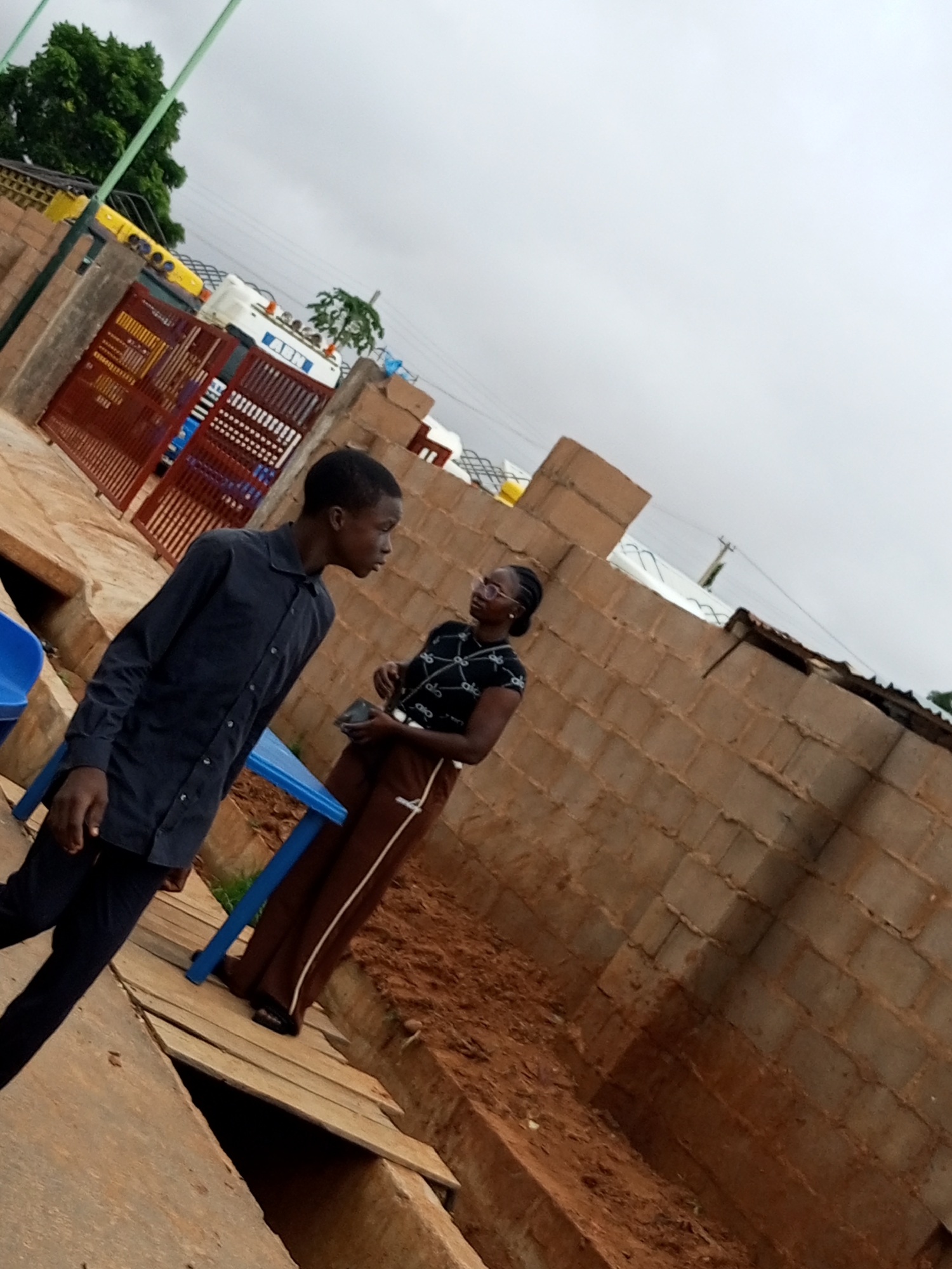 The image shows two individuals standing outdoors in what appears to be Garko, Nigeria. In the foreground on the left, a young male is walking towards the left side of the frame, wearing a dark long-sleeved shirt and dark pants. To his right, a young female is standing, facing towards the right of the frame. She is wearing glasses, a black shirt with white patterns, and brown pants. She is holding a dark object in her hands. Behind them, there is a wall constructed from light brown bricks. To the left of the individuals, a portion of a red metal gate is visible, and behind that, there are some yellow structures and a white structure with the letters "ARN" visible. The ground is made of dirt and some wooden planks forming a walkway. The sky is overcast. The scene is captured at an angle, with the ground appearing to tilt upwards from left to right.