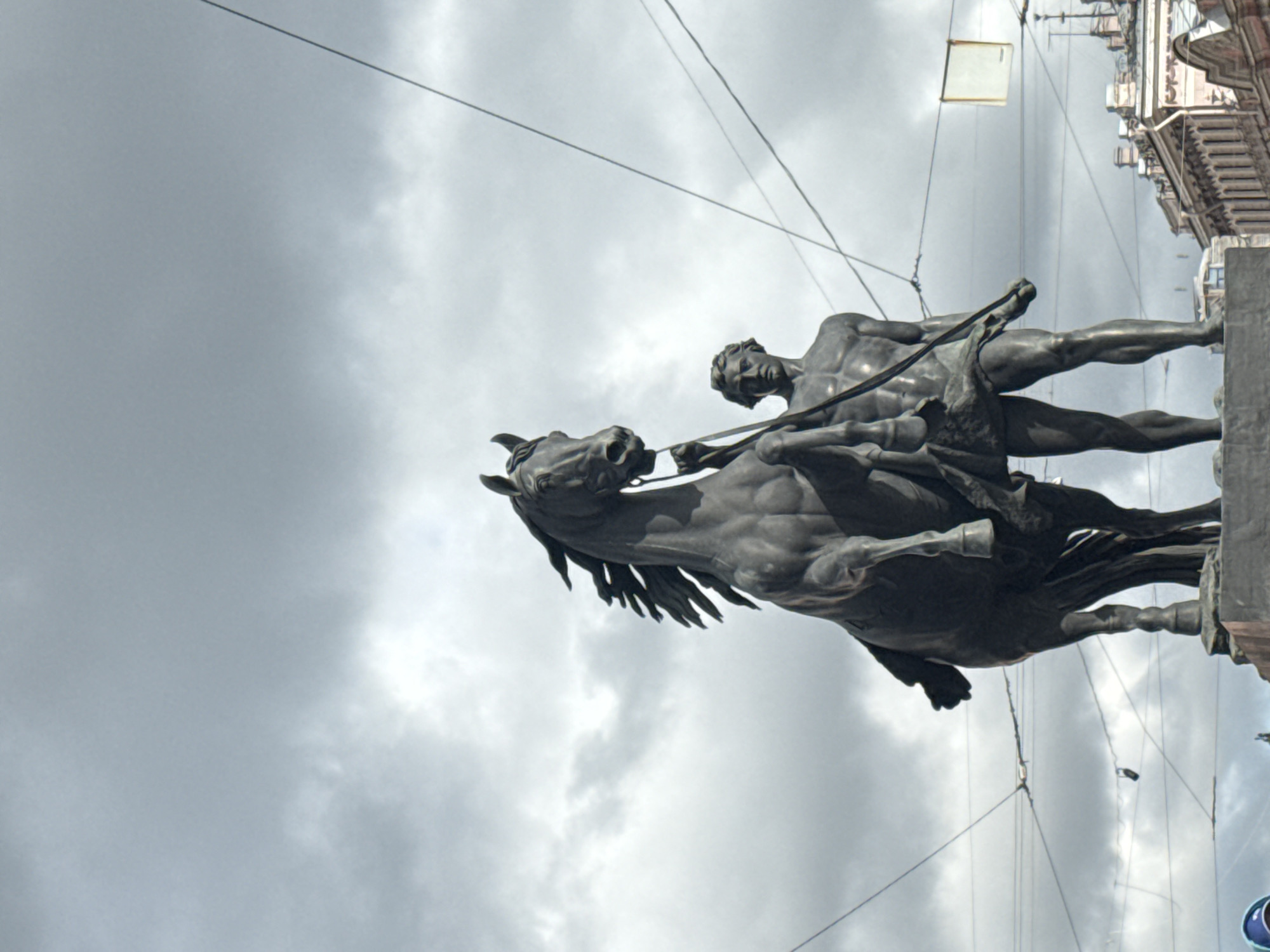 The image depicts a statue of a man riding a horse, prominently featuring muscular anatomy of both the rider and the horse. This is a statue in St. Petersburg, Russia. The sculpture is rendered in dark metal, likely bronze. Overhead power lines traverse the scene. The sky is overcast with gray clouds. A building is also visible in the background.