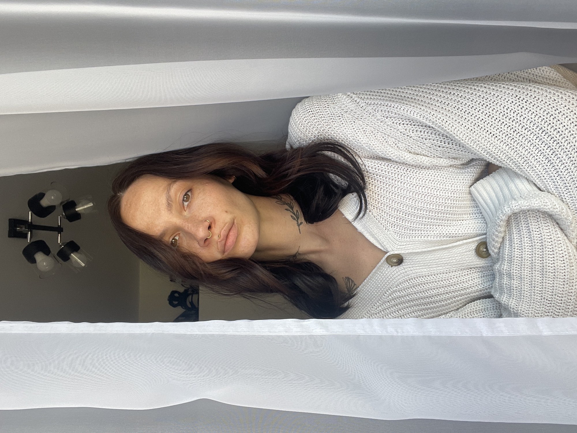 A woman with long brown hair and a white cardigan is positioned in front of a white, translucent curtain. The background includes a wall with a chandelier-like light fixture. No information indicates this photo was taken in Izhevsk, Russia.