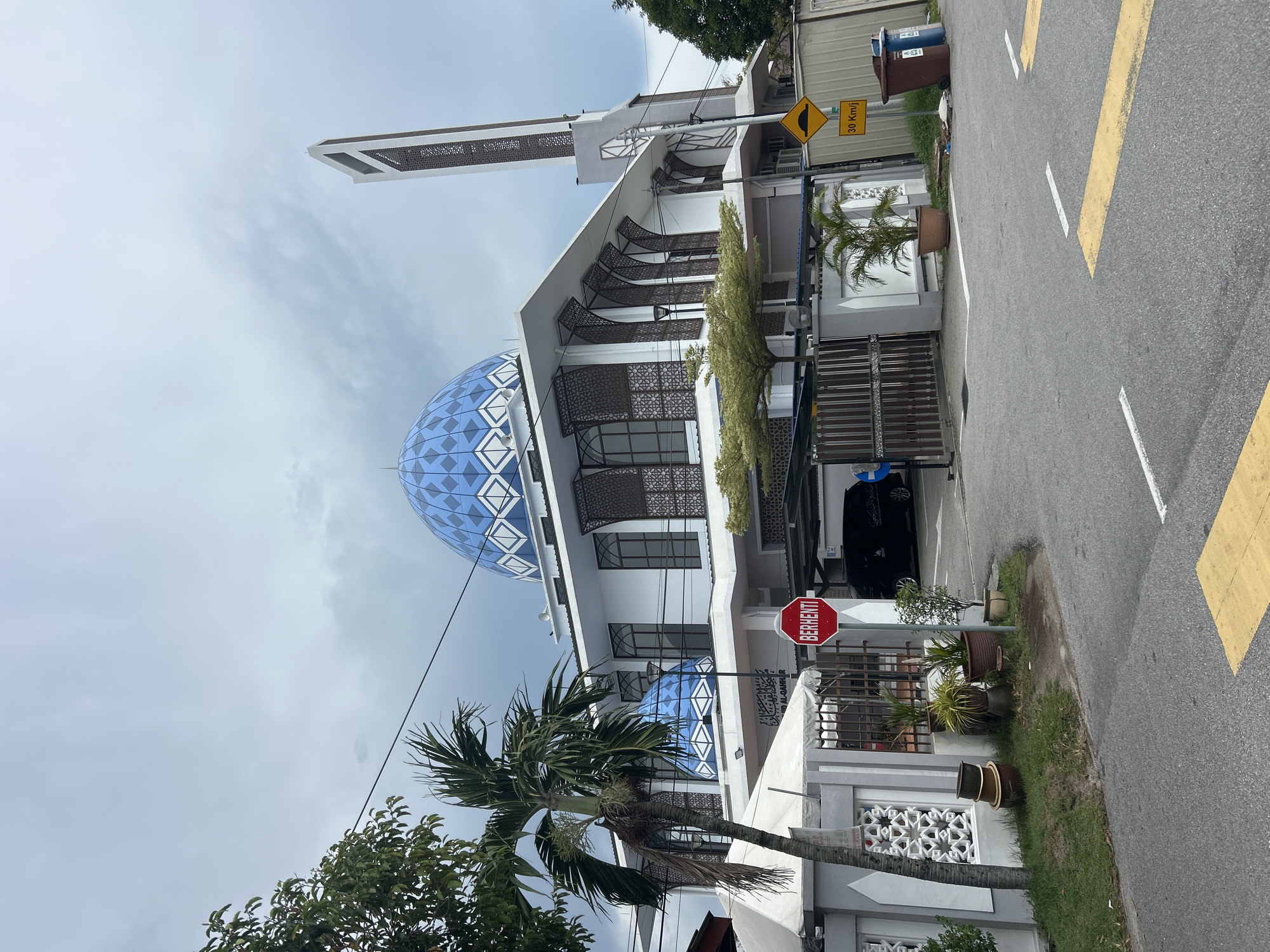 The image shows a mosque in George Town, Malaysia. The mosque has a large blue dome and a tall minaret. The building is white with intricate decorative screens. In front of the mosque, there is a street with road markings and a speed bump sign indicating 30 Km/h. A red stop sign with the word "BERHENTI" is visible. There are palm trees, potted plants, and a tent structure on the grounds. A black car is parked near the entrance. The sky is cloudy.