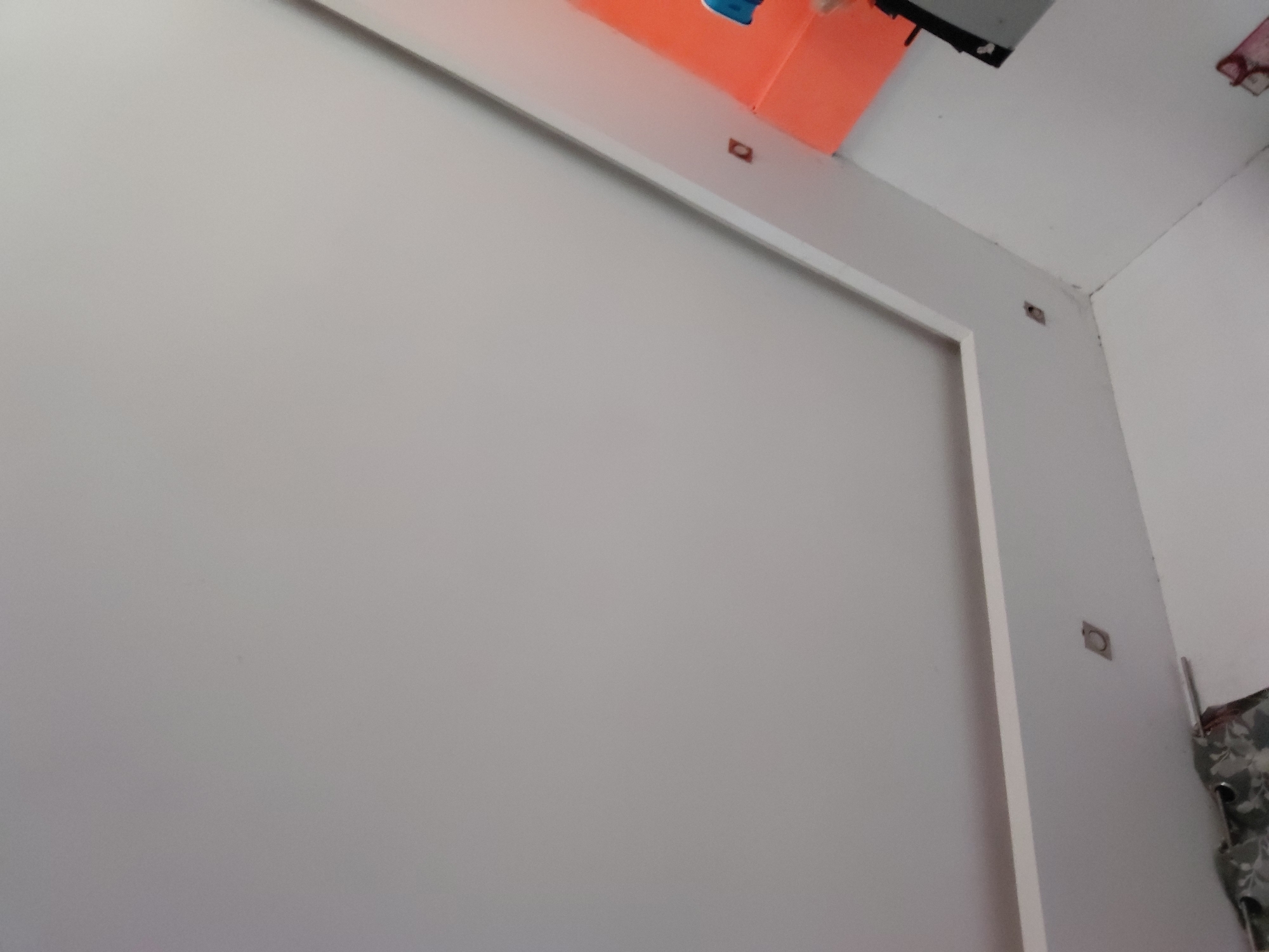 The image provides an interior view of a room, primarily focusing on a light grey wall featuring white architectural trim. This trim forms a large, incomplete rectangular frame on the wall. Several small, dark, square-shaped cutouts or depressions are visible within the white trim and on the adjacent wall surface, suggesting points for fasteners or fixtures.
In the upper left of the frame, an orange object is visible, possibly a shelf, a box, or part of a ceiling structure, with a small blue item attached to it. The top right corner reveals a wall intersection and a white ceiling, where some dark lines or minor imperfections are apparent. On the far right edge, a piece of patterned fabric, in shades of dark grey and white, is partially seen, likely a curtain or textile.
The scene appears to be an indoor setting, possibly a residential or commercial space. There are no individuals present, nor any specific activities taking place. The overall impression is of a static room detail, potentially during or after construction or renovation. No visible text is present in the image. The location context is Santa Ana, Venezuela.