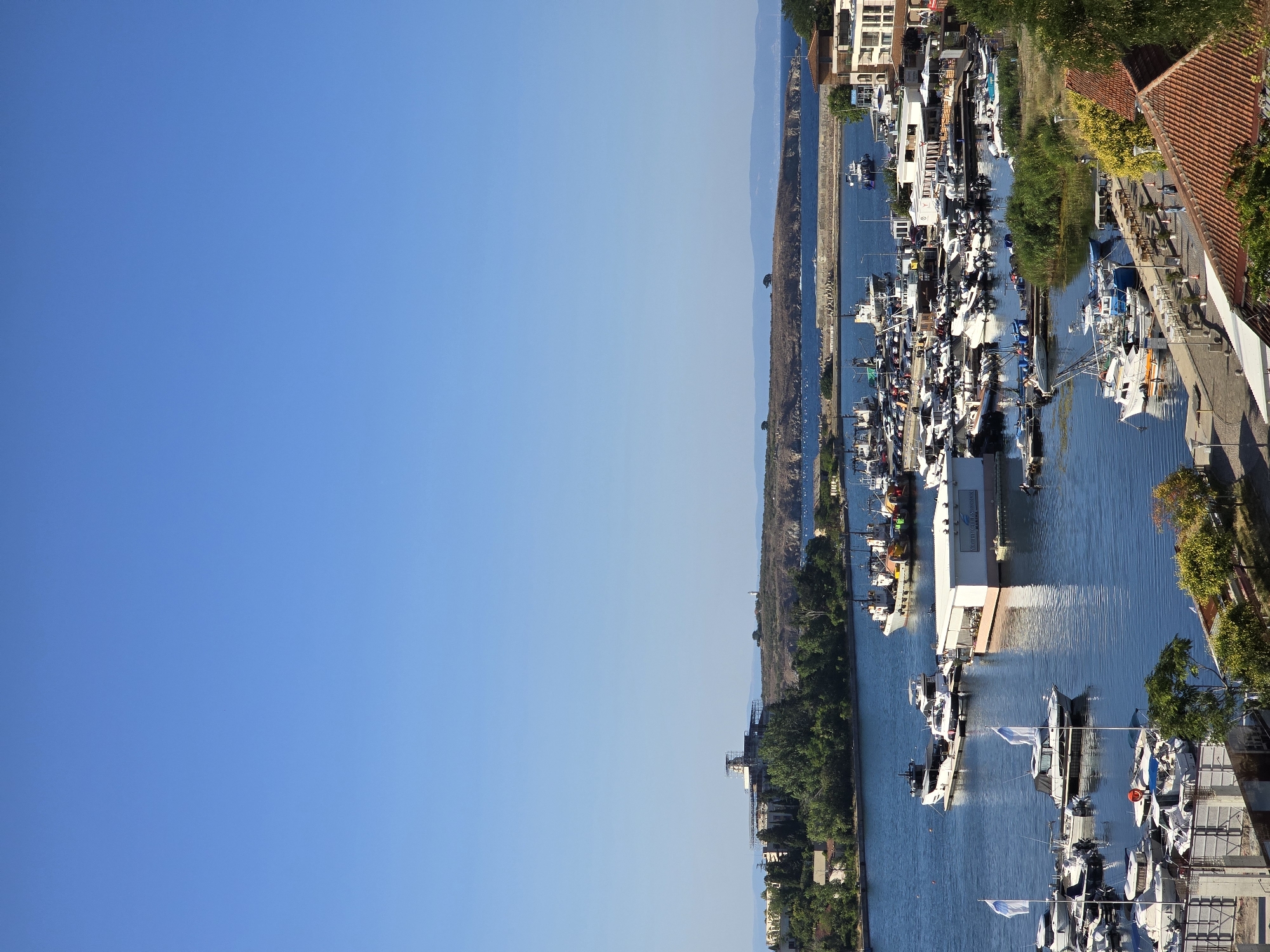 This is a high-angle, vertical, photographic shot of the Sozopol marina in Sozopol, Bulgaria. The sky is a clear, vibrant blue, taking up the top half of the image. Below the sky is a wide expanse of calm, blue water, which is the marina itself. Numerous boats of various sizes are docked or anchored in the water. These include small pleasure craft, larger yachts, and several fishing vessels. Some of the boats are white, while others have colored hulls and decks.
Along the edge of the marina are buildings. On the left side of the water, there is a cluster of buildings and trees, with a larger building structure on a distant hill, possibly a resort or historical site, with scaffolding around its upper portion. On the right side of the marina, there are more modern buildings, some with balconies and outdoor seating areas. Further back, a peninsula juts out into the sea, with sparse vegetation and a lighthouse visible on its tip.
The foreground shows a waterfront promenade with people walking and sitting at outdoor cafes. There are also more docked boats here, and some with flags. Trees and flowering bushes add greenery to the scene. The architecture includes buildings with red-tiled roofs. The overall impression is a sunny, peaceful day at a coastal town.