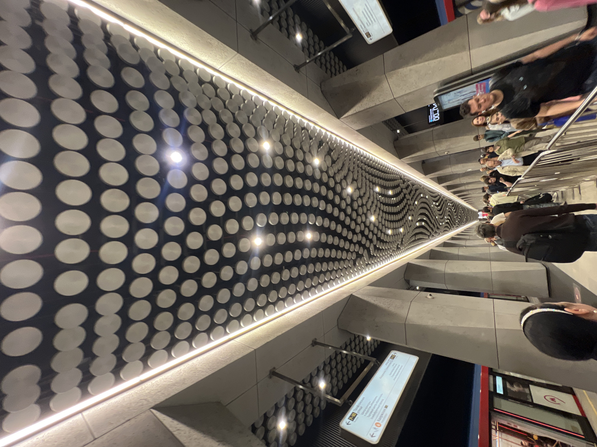 A metro station in Moscow, Russia, features a distinctive ceiling with a repeating pattern of circular indentations on a dark background, illuminated by long strip lights along the edges. The station has large, angled concrete columns supporting the ceiling structure. A modern metro train with red and blue accents is visible on the tracks to the left. Several people are walking along the platform, some are standing in line, and others are in the process of boarding or disembarking the train. Signage is visible on the platform, indicating directions and station names in Russian. The overall impression is one of a contemporary and well-lit urban transportation hub.