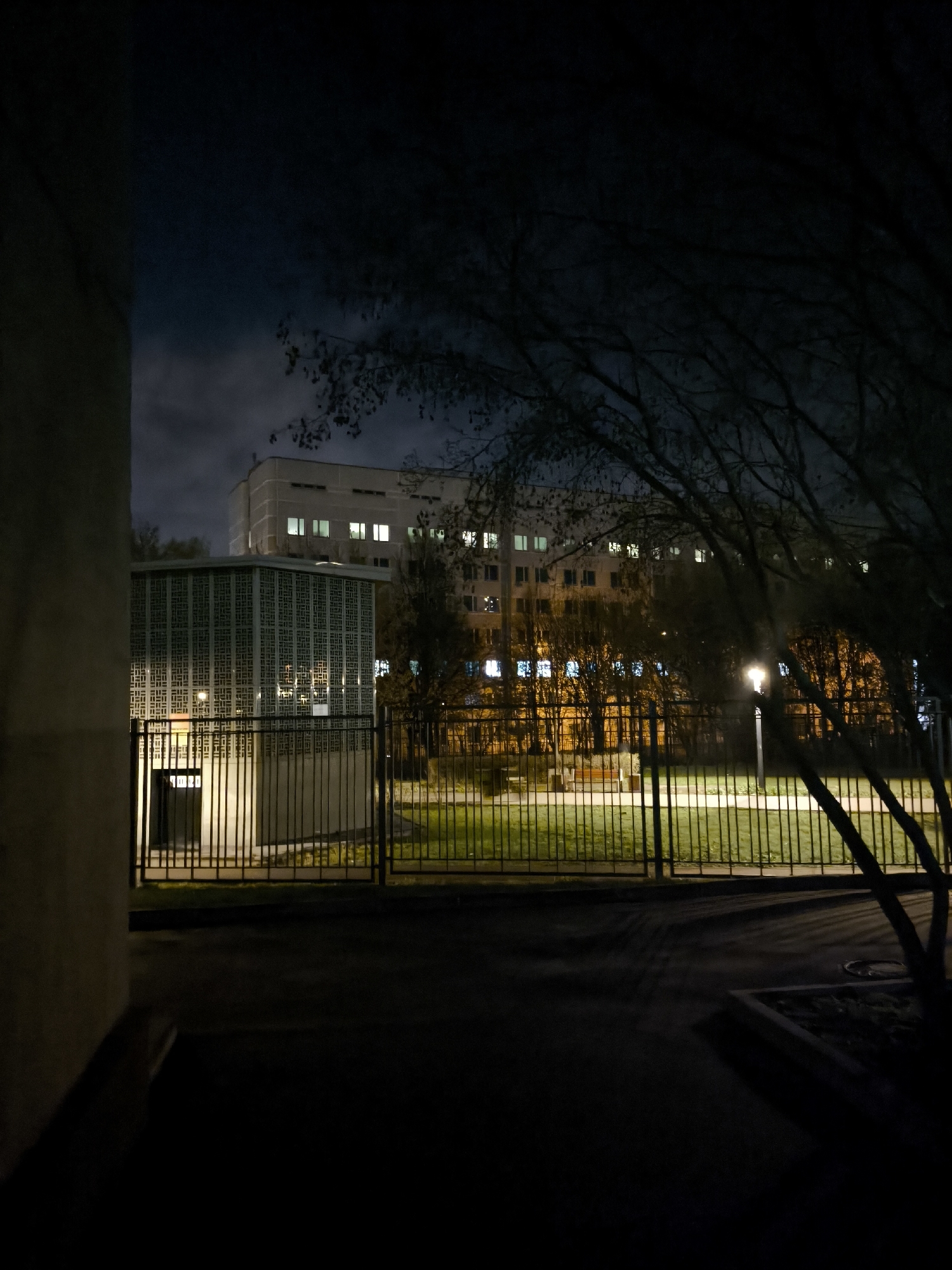 The image depicts a nighttime scene in Moscow, Russia. A building with illuminated windows is visible in the background. In the foreground, there is a fence, a smaller structure with a decorative facade, a tree with bare branches, and a paved area. The scene is lit by streetlights. No people are visible in the image.