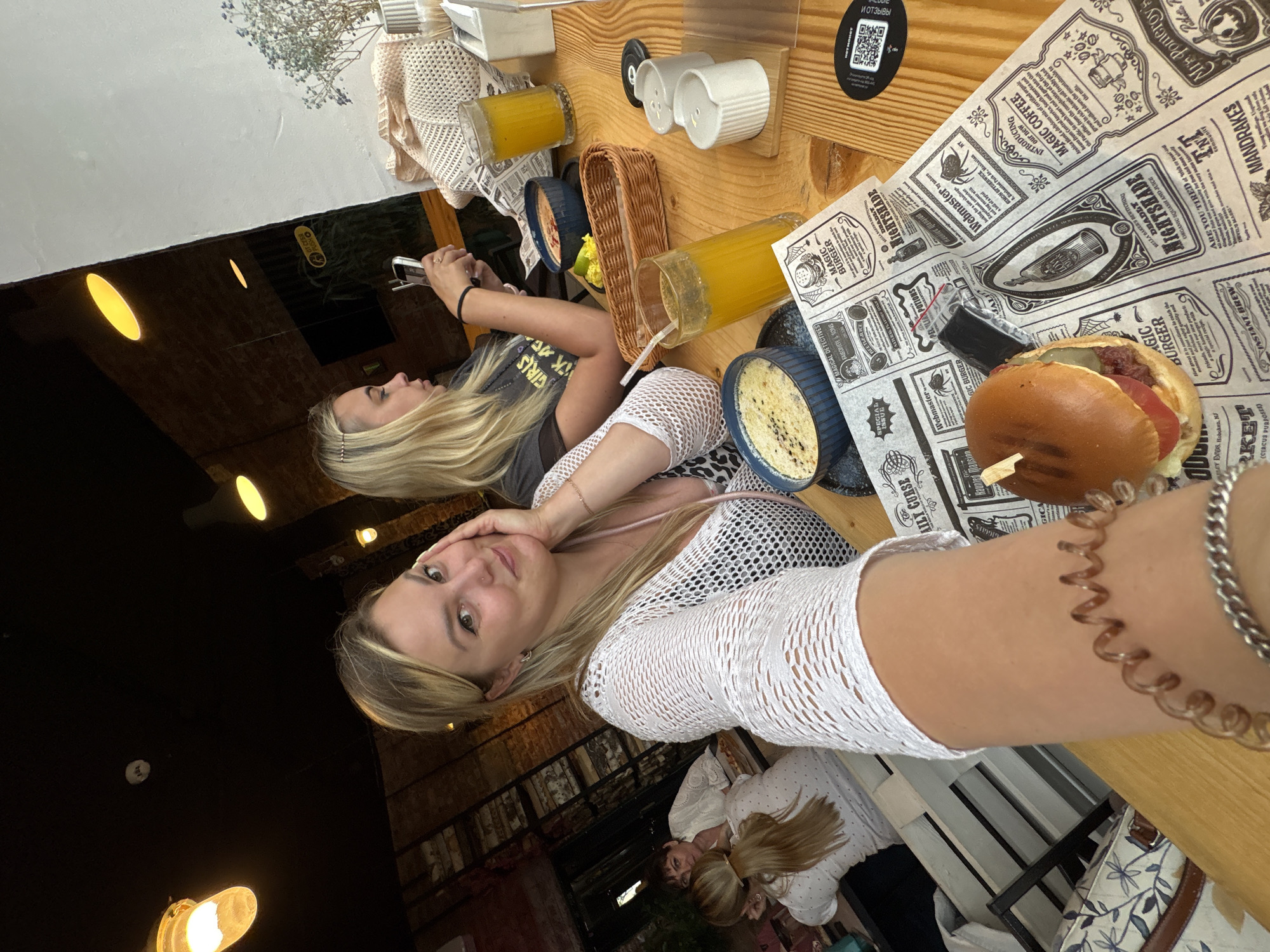 The image depicts a group of people dining at an indoor restaurant. In the foreground, a woman is taking a selfie, smiling and holding her hand to her face. She is wearing a white mesh cardigan over a black and white patterned top. A hamburger and a bowl of food are on the table in front of her, along with a glass of orange juice and a basket. Behind her, another woman with blonde hair is looking at her phone. Further in the background, two other women are seated at a table, one wearing a white top. The restaurant appears to have a rustic ambiance, with exposed brick walls and wooden tables. The ceiling has industrial-style lighting. The image does not provide any specific details about the city of Chaikovsky, Russia.