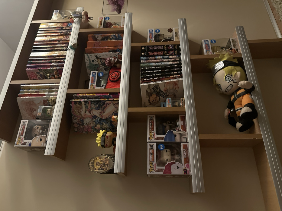The image depicts an interior scene, specifically a multi-tiered wooden bookshelf mounted against a light beige wall in a location within the city of Plovdiv, Bulgaria. The bookshelf features four visible shelves, each with a silver-colored horizontal bar along its front edge.
**Top Shelf:** Contains several Funko Pop figures in their original packaging, including one identifiable as "POP! ANIMATION 1132" (likely Master Roshi) and another partially visible. A smaller, green-haired Funko Pop figure is placed in front. To the right, numerous manga volumes with colorful spines are arranged vertically, some appearing to be from the *One Piece* series. A small, brown, turtle-like figurine sits near the manga. A hanging decorative item is suspended from the top right section of the shelf.
**Second Shelf (from top):** Features a rectangular, metallic-framed religious icon, likely depicting the Virgin Mary and Child, positioned on the far left. Next to it stands a Funko Pop figure of Naruto Uzumaki. To the right, a large, thick manga or art book with intricate, colorful cover art is prominently displayed alongside other manga volumes. A red, crocheted spiral object and a small, brown Funko Pop figure (possibly Gaara) are placed in front of some books. A partially visible Funko Pop box with "1134" is also present. A framed image with purple floral elements is affixed to the wall on the far right.
**Third Shelf (from top):** Displays two Funko Pop boxes from the "HUNTER x HUNTER" series: "652 HISOKA" and "1134 KITE," both bearing a "34.99" price tag. To their right, a collection of manga volumes is arranged, with "DEADMAN WONDERLAND Vol. 1" and several "Death Note" volumes clearly visible. A small figurine, possibly another Naruto character, is placed in front of these books. On the far right, a Funko Pop box labeled "932 ITACHI" is present.
**Bottom Shelf:** A large plush toy of Naruto Uzumaki, wearing his signature orange and black jumpsuit and a headband, lies horizontally on its back, facing right. A partially visible Funko Pop box with a "34.99" price tag is visible behind the plush toy on the right.
The overall lighting is warm, originating from an unseen source above and to the left, casting soft shadows. No people are present in the scene. The items are neatly arranged, indicating a curated display of collectibles and literature.
