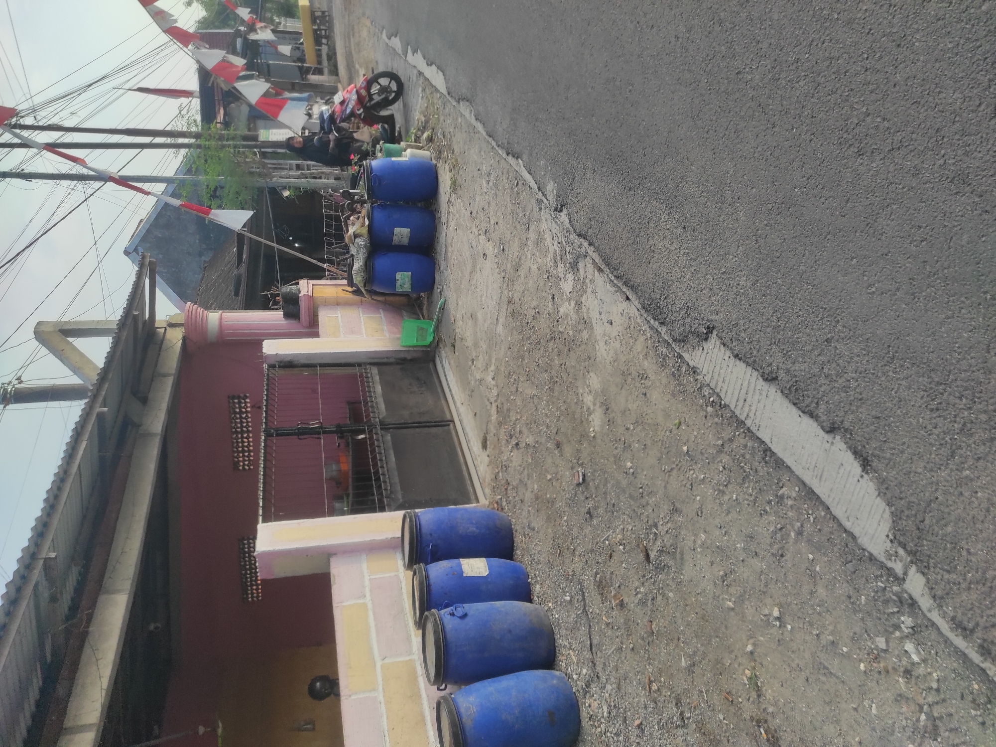 In Klepek, Indonesia, this outdoor scene captures a residential or small commercial area with a focus on utility. A pink building with a corrugated metal roof dominates the left side, its facade interrupted by a metal gate. In front of the building, four large blue plastic barrels stand in a row, suggesting storage or waste management. To the right, a person wearing a dark hijab is seated on a red motorcycle. Indonesian flags, red and white, are strung across poles and wires, indicating a possible celebration or national event. The ground is a mix of gravel and asphalt, with a concrete curb running alongside the road. The overall impression is one of everyday life in a modest Indonesian community, with practical elements like barrels and a motorcycle juxtaposed with patriotic decorations.