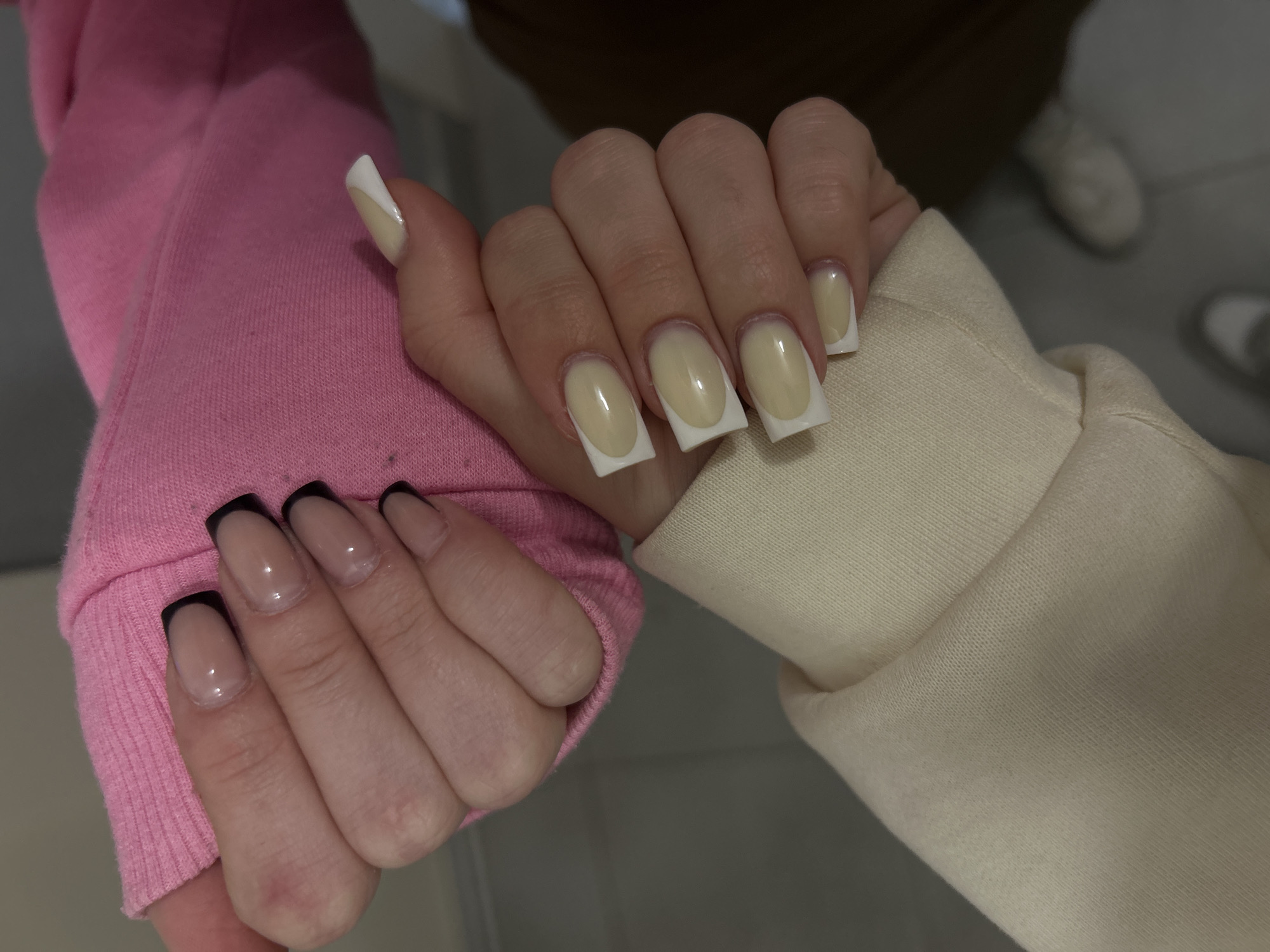The image captures a close-up view of two hands, likely belonging to two different people, set against a softly blurred indoor background. The hands are positioned side-by-side, with the left hand slightly overlapping the right, showcasing contrasting nail art and clothing.
The hand on the left is adorned with a bright pink, ribbed long-sleeved garment, possibly a sweatshirt or hoodie. Its nails are of medium length, shaped in a rounded square, featuring a translucent nude base and distinct black French tips.
The hand on the right wears a cream or light beige long-sleeved garment with a smoother texture. This hand displays longer, square-shaped nails with a creamy or pale yellow base coat and crisp white French tips. Both sets of nails are glossy and well-maintained.
The activity appears to be a deliberate pose to highlight the different nail designs and clothing styles. The background is out of focus, showing indistinct shapes that suggest an indoor setting, possibly with a light-colored floor and blurred footwear in the distance. The lighting is soft and even, suggesting an indoor environment. There is no visible text in the image.
