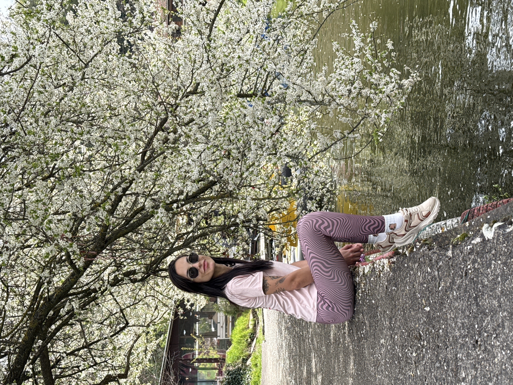 A woman is sitting on a concrete edge near water, under a tree covered in white blossoms. She is wearing sunglasses, a light pink t-shirt, and patterned leggings. Her sneakers are visible, and a tattoo is visible on her arm. The location is in Pleven, Bulgaria, near a body of water, with vegetation and buildings visible in the background. The camera angle is from below, looking up at the subject.