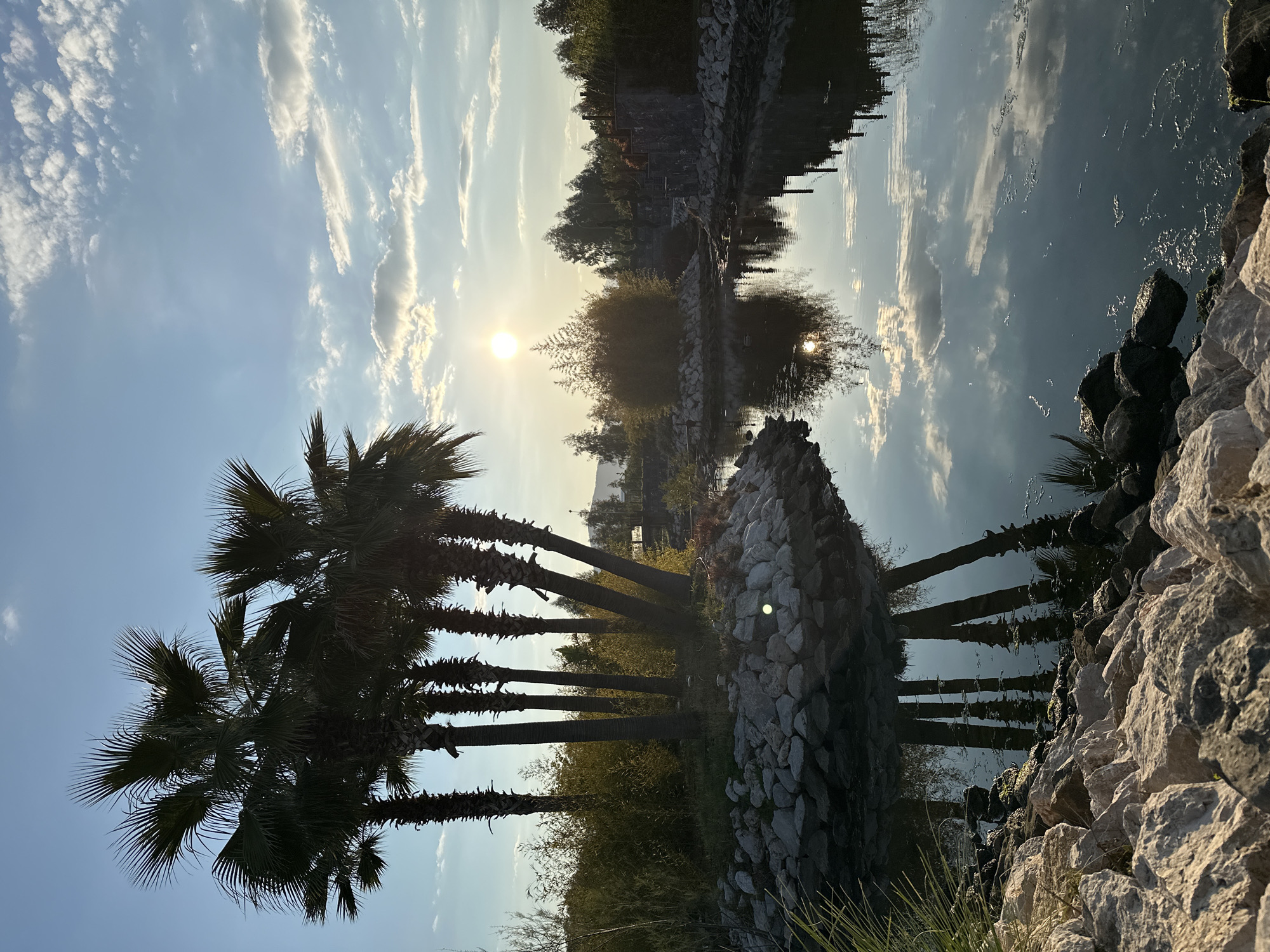 The image captures a tranquil scene in Fethiye, Turkey, featuring a body of water reflecting the sky. The sky is partially cloudy with the sun visible, creating a bright glare. Palm trees line the foreground on the left, contrasting against the lighter sky. A stone structure or embankment runs along the water's edge, reflecting in the water. Rocks and vegetation are present along the bank. The image is oriented in a landscape format.