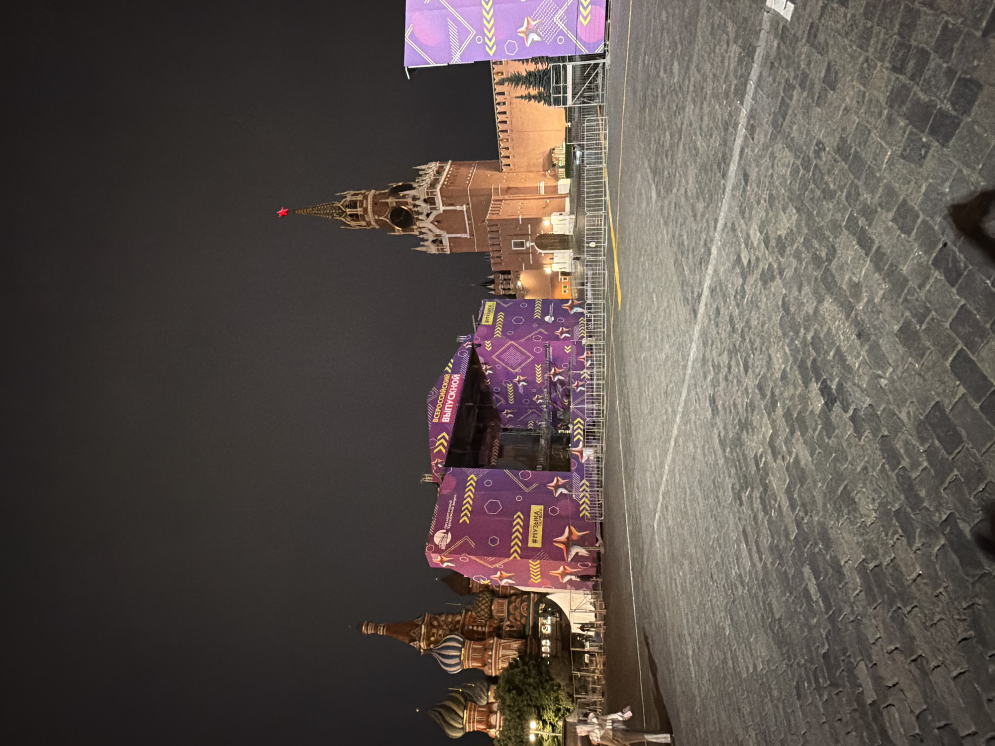 An exterior nighttime wide shot captures a section of Red Square in Moscow, Russia. In the center of the frame, a large, temporary stage structure, adorned with purple and yellow geometric patterns and stars, is erected. Text in Russian, including "ВСЕРОССИЙСКИЙ ВЫПУСКНОЙ" (All-Russian Graduation) and "#МУЗЫКА ПЕРВОГО" (#MusicOfTheFirst), is visible on the stage's facade. To the left of the stage, the iconic multi-colored domes of Saint Basil's Cathedral are partially illuminated against the dark sky. To the right of the stage, the prominent red brick Spasskaya Tower of the Moscow Kremlin stands tall, topped with a glowing red star. The ground in the foreground is paved with dark cobblestones, and metal barricades are visible, suggesting an event is being prepared or is underway. A single person, a woman wearing a light-colored jacket and dark pants, is walking on the cobblestones to the left of the stage. The overall atmosphere is festive, with the stage lighting and the illuminated Kremlin and cathedral contributing to the scene.