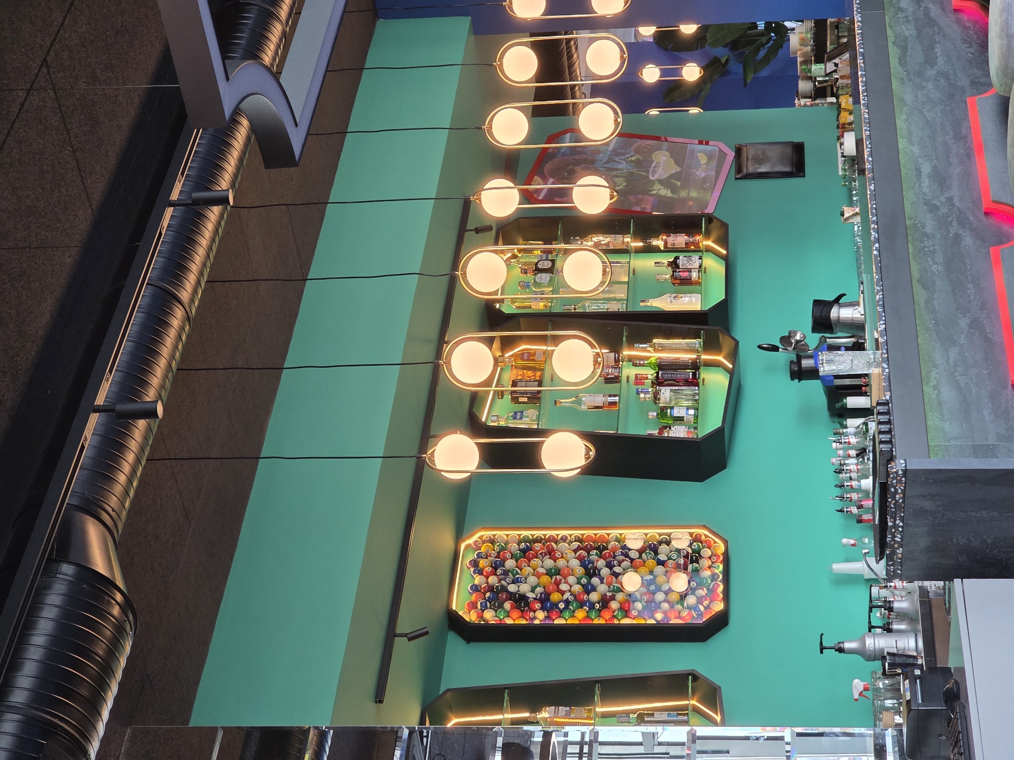 The image depicts the interior of a bar, possibly located in Varna, Bulgaria, though this cannot be confirmed definitively from the visual data. The scene features a vertical wall painted in shades of teal. Mounted on the wall are several illuminated display cases. One contains a collection of billiard balls. Others hold assorted liquor bottles. Above these are two neon-illuminated display cases, one of which contains a framed picture. A row of modern, spherical pendant lights hang down from the ceiling. A section of the bar top is visible, equipped with beverage dispensers and other bar equipment. An exposed industrial-style ventilation duct is visible on the left side of the image.
