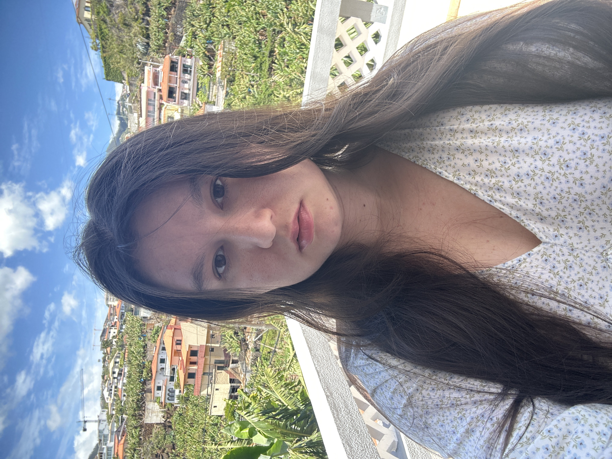 A young woman with long, dark hair is posing for a selfie. She is wearing a light-colored, floral-patterned top. The background shows a cityscape of Câmara de Lobos, Portugal, including buildings and vegetation. The sky is blue with some clouds. A white balcony railing is partially visible.