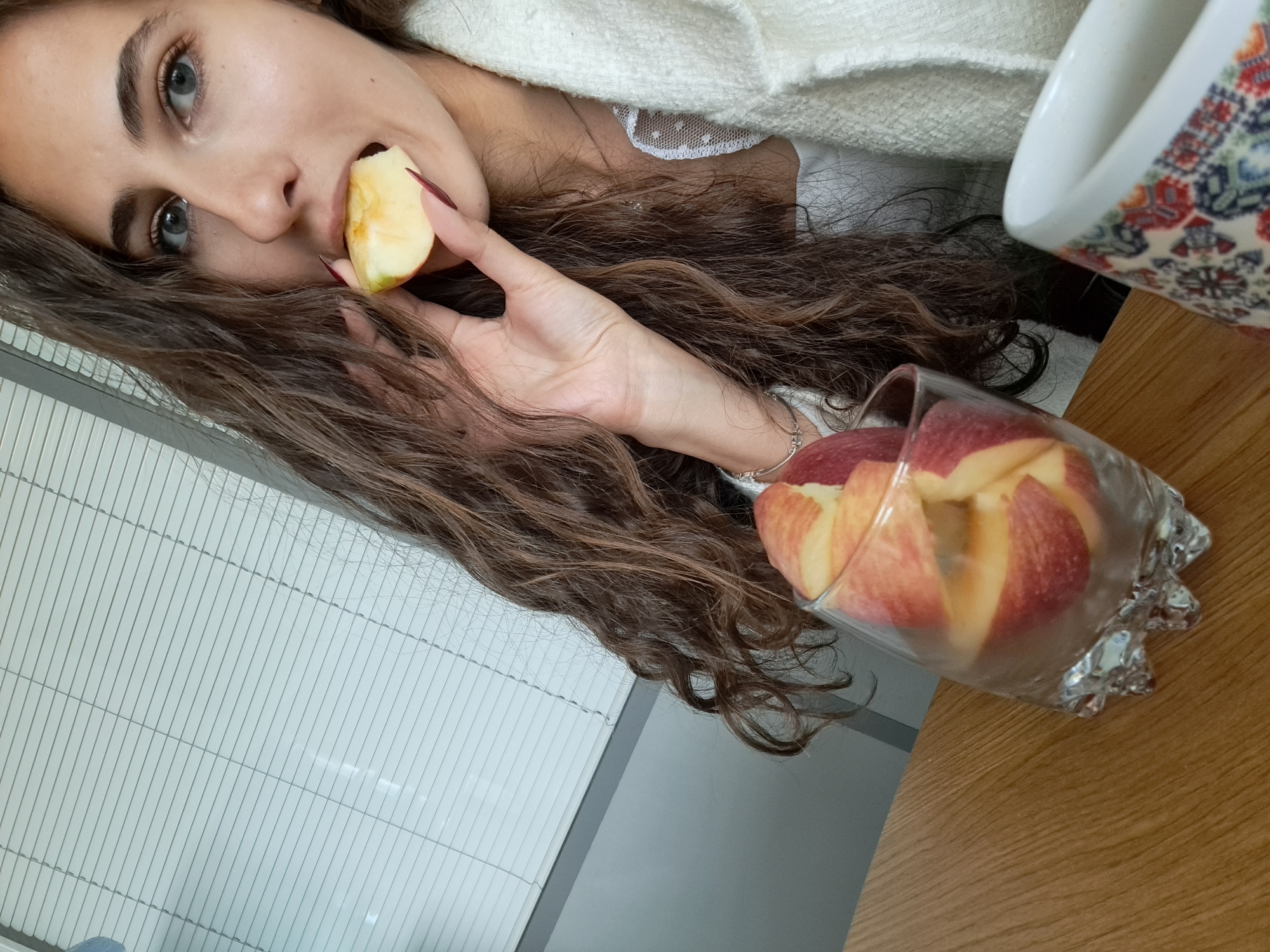 A woman with long, brown, curly hair is eating an apple slice. She is holding the apple slice in her right hand, bringing it to her mouth. A glass filled with apple slices is on a wooden table next to her. A white ceramic mug with a colorful pattern is also visible on the table. She is wearing a white top with lace details, covered by a white sweater. The background shows a window with blinds. The location cannot be determined solely from the image, as there is no visible landmark to indicate it is Sofia, Bulgaria.