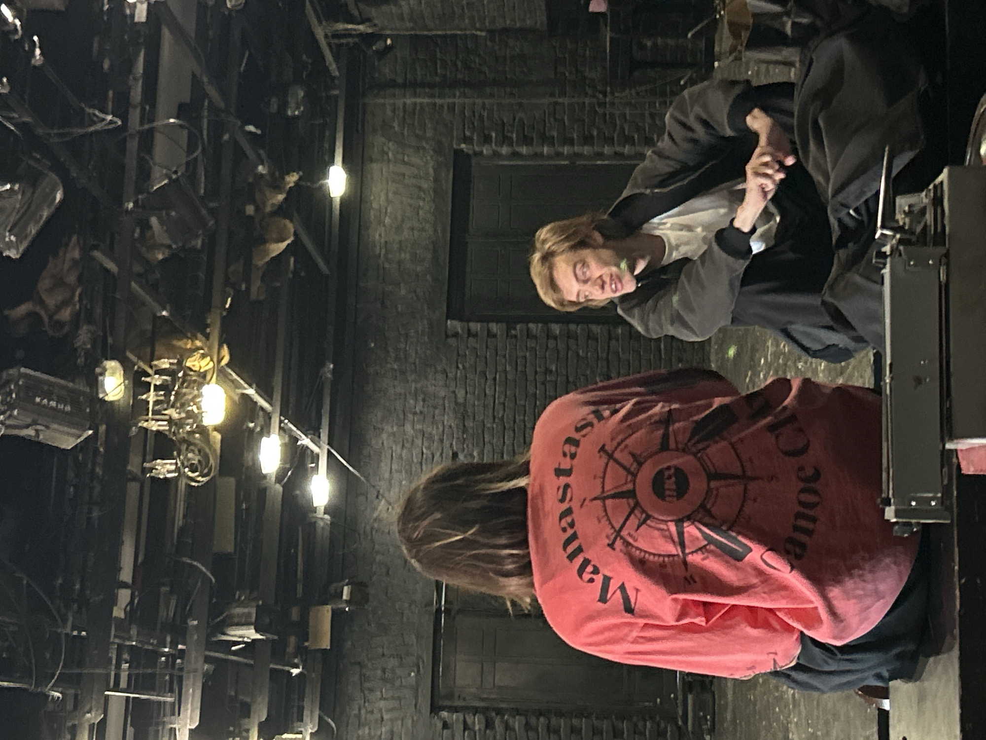 The image shows two individuals in an indoor, industrial-looking setting. The person in the foreground is viewed from behind, wearing a long-sleeved pink shirt with a large compass design printed on the back. Text on the shirt reads "Manastash Canoe Club". The person has long, light brown hair. The second individual, in the background, is facing towards the camera and appears to be a young person with blonde hair. They are wearing a dark hooded jacket over a white t-shirt and dark pants. Their head is tilted, and they are looking down, with their mouth slightly open, suggesting they might be speaking or reacting to something.
The setting appears to be a workshop or storage area, with various metal structures, pipes, and what look like salvaged or industrial equipment in the background. Several light fixtures are visible, casting a warm glow. The walls are made of dark brick. The lighting suggests it is either evening or an interior space with artificial lighting. There is no indication of the weather. The overall mood is casual, with the subjects seemingly engaged in a conversation or interaction. The image is oriented vertically, with the bottom of the frame at the top.