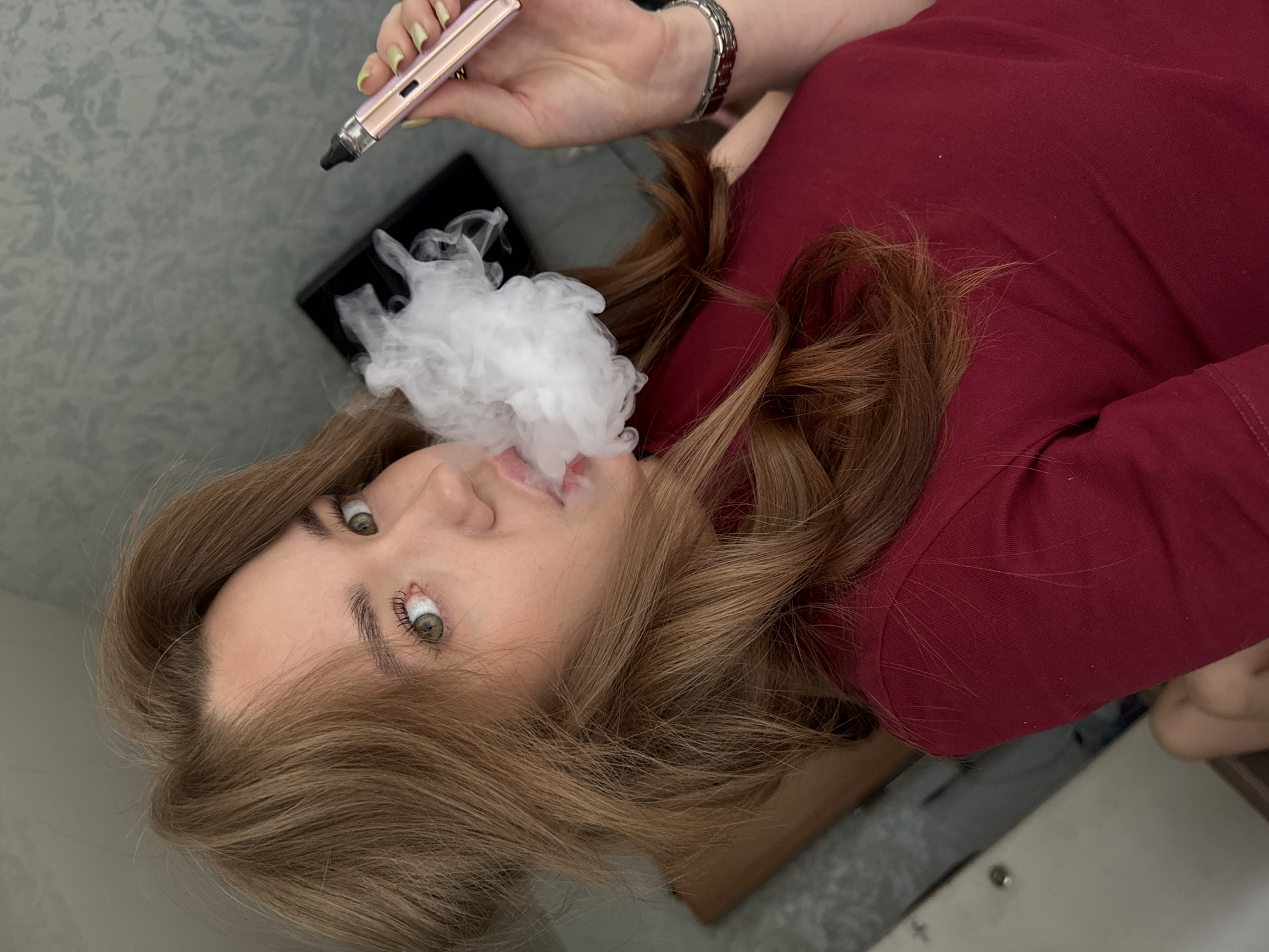 In this close-up shot, a young woman with shoulder-length brown hair exhales a cloud of white vapor. She is wearing a dark red, long-sleeved shirt and has green eyes. She is holding a rose gold vape pen in her right hand, and a silver watch is visible on her left wrist. The background appears to be an indoor setting, possibly a room with a textured gray wall and a dark object, perhaps a desk or laptop, behind her. The image does not contain any information that would indicate its location in Chaikovsky, Russia.